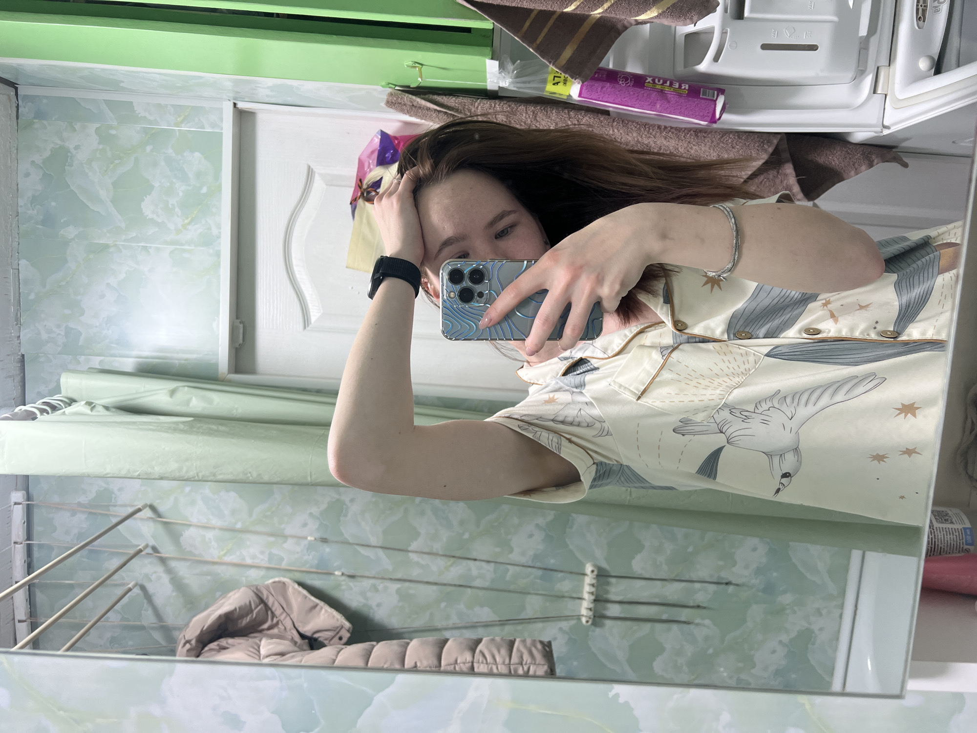The image is a mirror selfie of a young woman, seen from the torso upwards, holding a smartphone to capture her reflection. The original image is oriented horizontally, with the woman's head facing left.
The woman has long, dark hair. She is wearing a light-colored button-up top, possibly pajamas, adorned with a pattern of gray/blue birds (resembling seagulls or cranes) and gold starbursts. On her left wrist, she wears a black digital smartwatch, and on her right, a silver-colored braided bracelet. Her face is partially obscured by the smartphone she holds, which has a silver or light blue patterned case and a multi-lens camera system.
The setting, visible in the mirror reflection, appears to be a domestic bathroom or utility room. The walls are covered with light green/aqua tiles featuring a mottled, cloud-like pattern. A section of a white door with a decorative panel is visible to the left of her head. Above the door and tiles is a light green shelf or cabinet, on which a brown striped towel and a purple rectangular box are placed. Due to the mirror reflection, the text on the purple box reads "XUJBOX" and "47g" backward.
In the lower part of the reflection, a wall-mounted clothes drying rack with multiple rods is visible, and a light brown/beige puffer jacket is draped over it. The room appears brightly lit, likely by indoor lighting. The overall impression is a functional, lived-in interior.