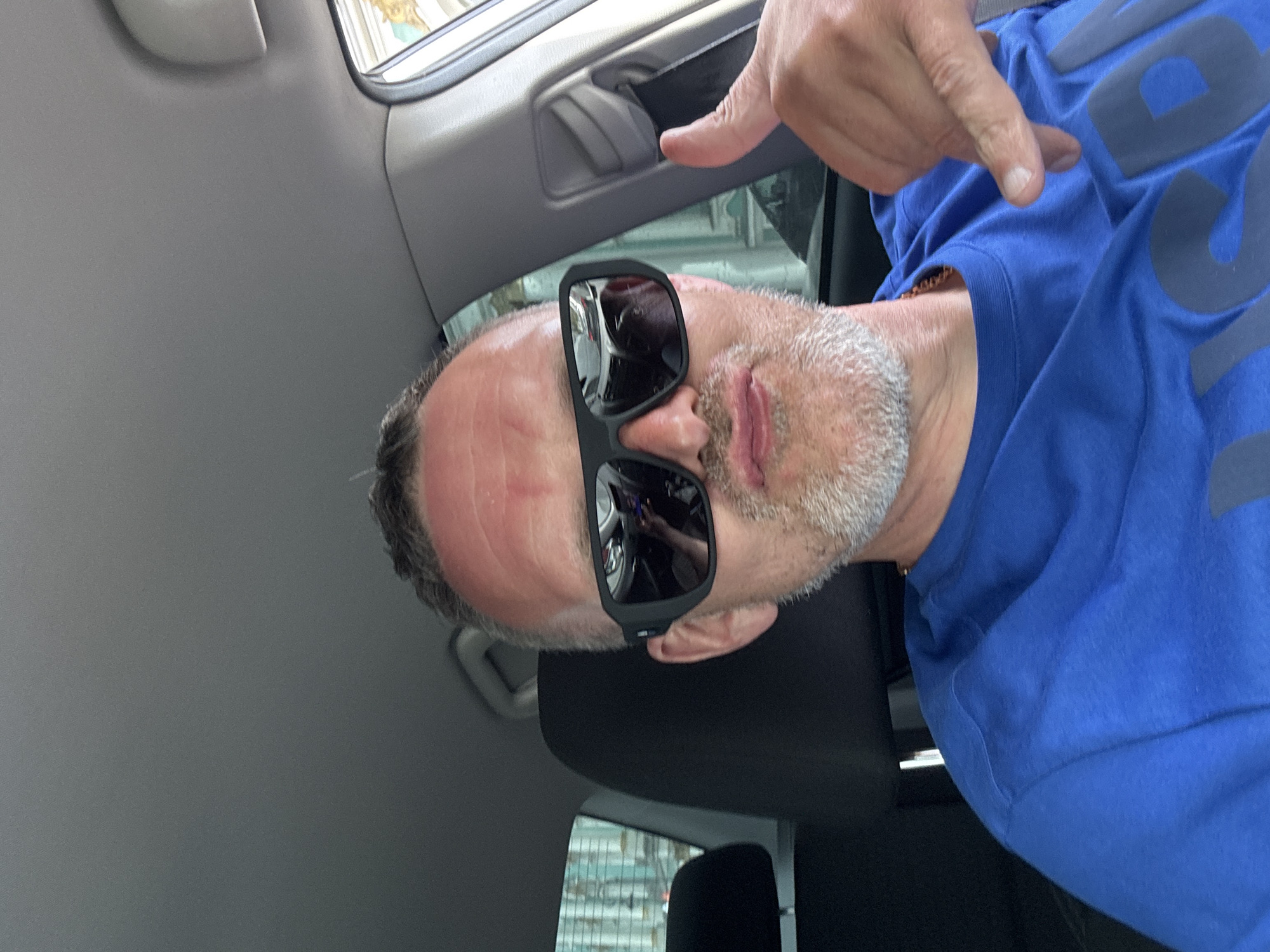 A middle-aged man wearing sunglasses and a blue shirt is taking a selfie inside a car. The image is oriented vertically, showing a close-up of his face and torso. His left hand is raised, pointing toward his chest. The background shows the car's interior and a blurred view of an exterior scene, possibly taken in Saint Petersburg, Russia, based on the architectural style and signs visible in the reflection of the sunglasses.