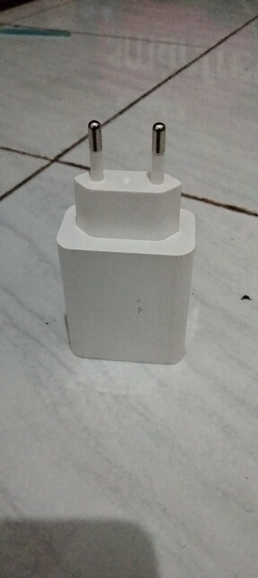 A white power adapter with two metal prongs is positioned on a tiled surface. The tiles are a light gray color with visible grout lines forming a grid pattern. The adapter has a two-tiered design, with a smaller upper section and a larger base. The prongs are metallic and extend from the top of the upper section. The scene appears to be indoors, possibly in a residential setting in Antirogo, Indonesia.