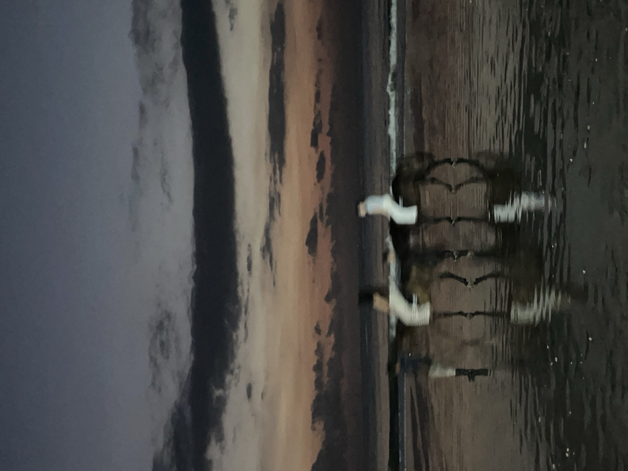 A vertically oriented image captures a serene twilight scene, split between a dramatic sky on the left and a reflective body of water on the right. The sky is filled with heavy, dark grey clouds, interspersed with streaks of soft orange and lighter grey, indicating either sunset or sunrise.
On the right side, a calm body of dark water extends horizontally, beautifully reflecting the sky's colors and the figures within it. Two individuals, appearing to be people, are positioned within this water. They are largely obscured by the water's surface and their pronounced reflections. Both individuals are wearing light-colored tops, which contrast with the darker hues of their lower bodies and the surrounding water. Their figures are elongated and somewhat distorted by the reflections, stretching horizontally across the water's surface.
The individuals appear to be standing or wading gently, creating a tranquil and almost abstract composition due to the low light and strong reflections. The scene evokes a contemplative and peaceful mood. There is no visible text.