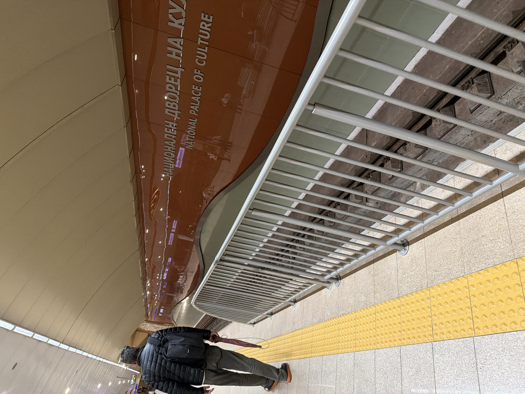 The image shows the National Palace of Culture metro station in Sofia, Bulgaria. The station's walls are decorated with the inscription "НАЦИОНАЛЕН ДВОРЕЦ НА КУЛТУРА" and "NATIONAL PALACE OF CULTURE." A man in a black jacket and gray pants stands on the platform, near a tactile paving intended to assist visually impaired individuals. He holds an umbrella. The platform is lined with a metal railing.