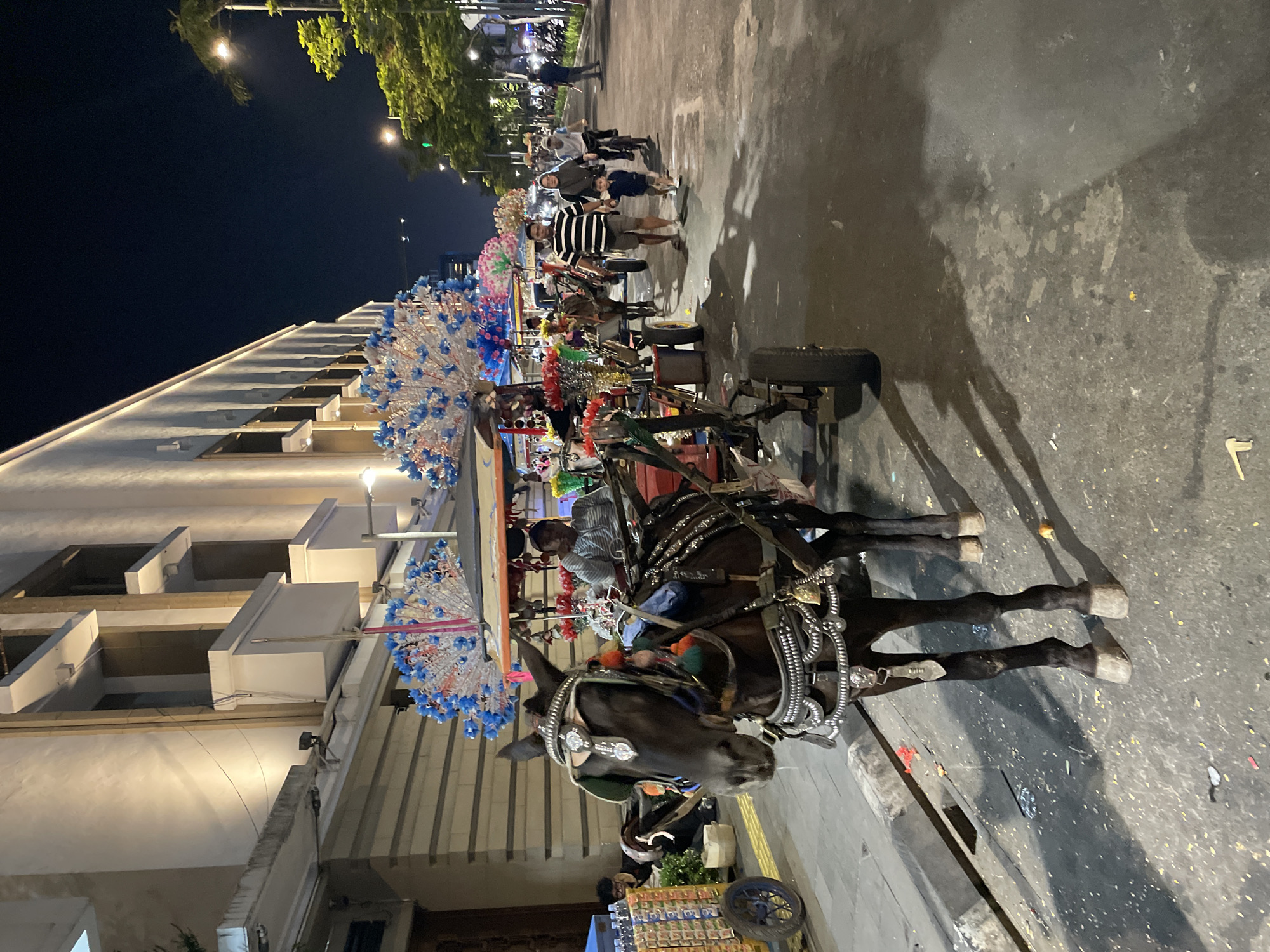 A horse-drawn carriage is the main subject, positioned in the foreground and angled to the left. The horse is dark brown and adorned with decorative silver bridles, reins, and girth, including a bell on the girth. The carriage itself is also decorated with colorful embellishments, possibly artificial flowers or feathers, in blue and white. A man, likely the driver, sits on the carriage, wearing a striped shirt and a cap. In the background, a building with a light-colored facade and illuminated windows lines the street. Several people are walking and cycling on the pavement and street. The scene appears to be in a city environment at night, with streetlights illuminating the area. Some decorative stands or carts are also visible along the street. Based on the context of horse-drawn carriages and street vendors, the location is likely Kota Tua (Old Town) in West Jakarta, Indonesia, a popular tourist area.