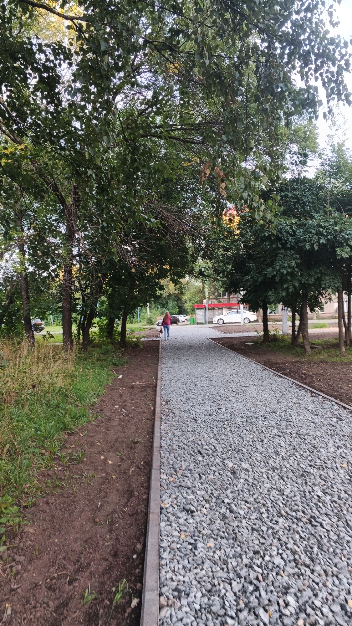 The image shows a gravel pathway with a dirt and grass area to its left. A person in a pink jacket and blue jeans is walking away from the camera on the pathway, seemingly pushing a stroller. In the background, there are trees, several cars, and a bus stop with a red canopy. The scene suggests a public park or residential area in Izhevsk, Russia, possibly during late summer or early autumn, given the foliage. The lighting indicates it's daytime, with overcast conditions.