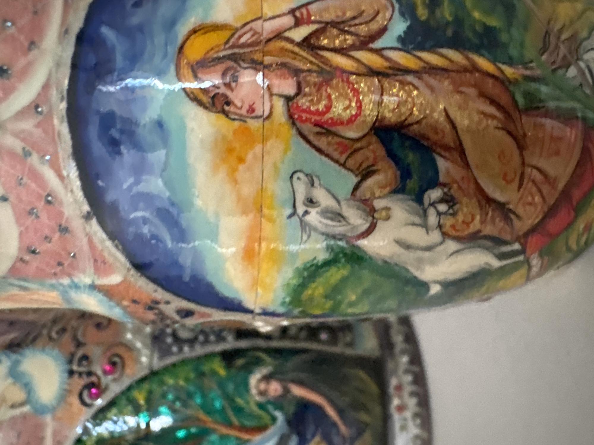 The image presents a close-up of a hand-painted decorative object, likely a traditional Eastern European or Russian folk art piece such as a Matryoshka doll or lacquer box, due to its style and curved, glossy surface. The central painting features a blonde-haired woman with a long braid, wearing a golden-brown garment with red accents and shimmering glitter. She is depicted interacting with a small white animal, possibly a goat or lamb, which gazes up at her.
The background of this scene transitions from a deep blue sky into warm yellow and orange hues, suggesting either sunrise or sunset, above a lush green landscape. A bright, horizontal light reflection spans across the center of the glossy surface.
A lower section of the object is visible on the left, revealing additional intricate patterns, including spirals and embedded pink rhinestones, along with a glimpse of another painted figure, possibly a dark-haired woman, against a vibrant, shimmering green and blue background. The overall aesthetic is rich in detail and color, indicative of a handcrafted item. There is no visible text in the image. While the location context is Puebla, Mexico, the artistic style strongly suggests an Eastern European origin for the depicted artwork.
