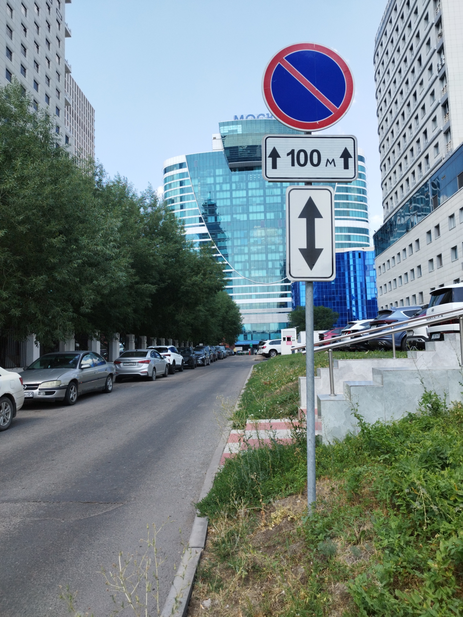 In Astana, Kazakhstan, on a clear day, a street scene unfolds with modern architecture and traffic signage. A row of parked cars lines the asphalt road, with a silver sedan prominently positioned on the left, followed by other vehicles, including a white SUV, a blue sedan, and a black car. Towering over the scene are contemporary buildings; on the left, a light-colored, multi-story structure with numerous windows stands tall. To its right, a striking blue glass skyscraper with an undulating facade dominates the skyline. On the far right, another large building, primarily white with a grid of windows, is visible.
In the foreground, a traffic signpost stands on the right side of the road. At the top, a circular red and blue sign with a diagonal red line indicates "No Stopping." Below it, a rectangular white sign displays "↑ 100 m ↑" with two upward-pointing arrows. Beneath this, another white rectangular sign features a single vertical double-headed arrow. Lush green trees with dense foliage line the street between the road and the buildings, creating a natural buffer. The ground near the signpost is covered with grass and small plants, with a concrete structure and steps to its right. The sky is a pale, cloudless blue.