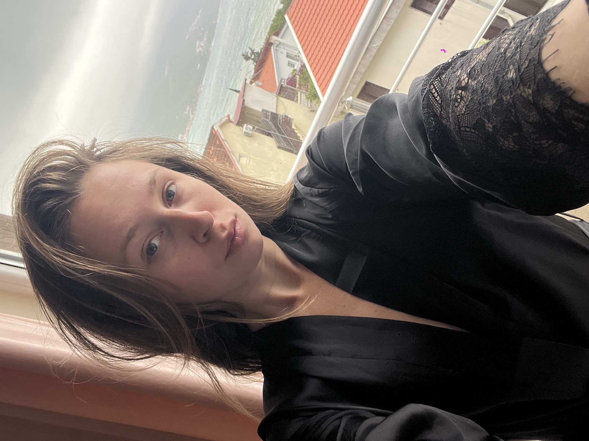 A woman with shoulder-length brown hair, wearing a black satin robe with lace sleeves, is taking a selfie on a balcony. The background shows a view of Bečići, Montenegro, with its characteristic red-tiled roofs and buildings facing the sea. The sky is overcast, suggesting a cloudy day. The balcony has a white railing.