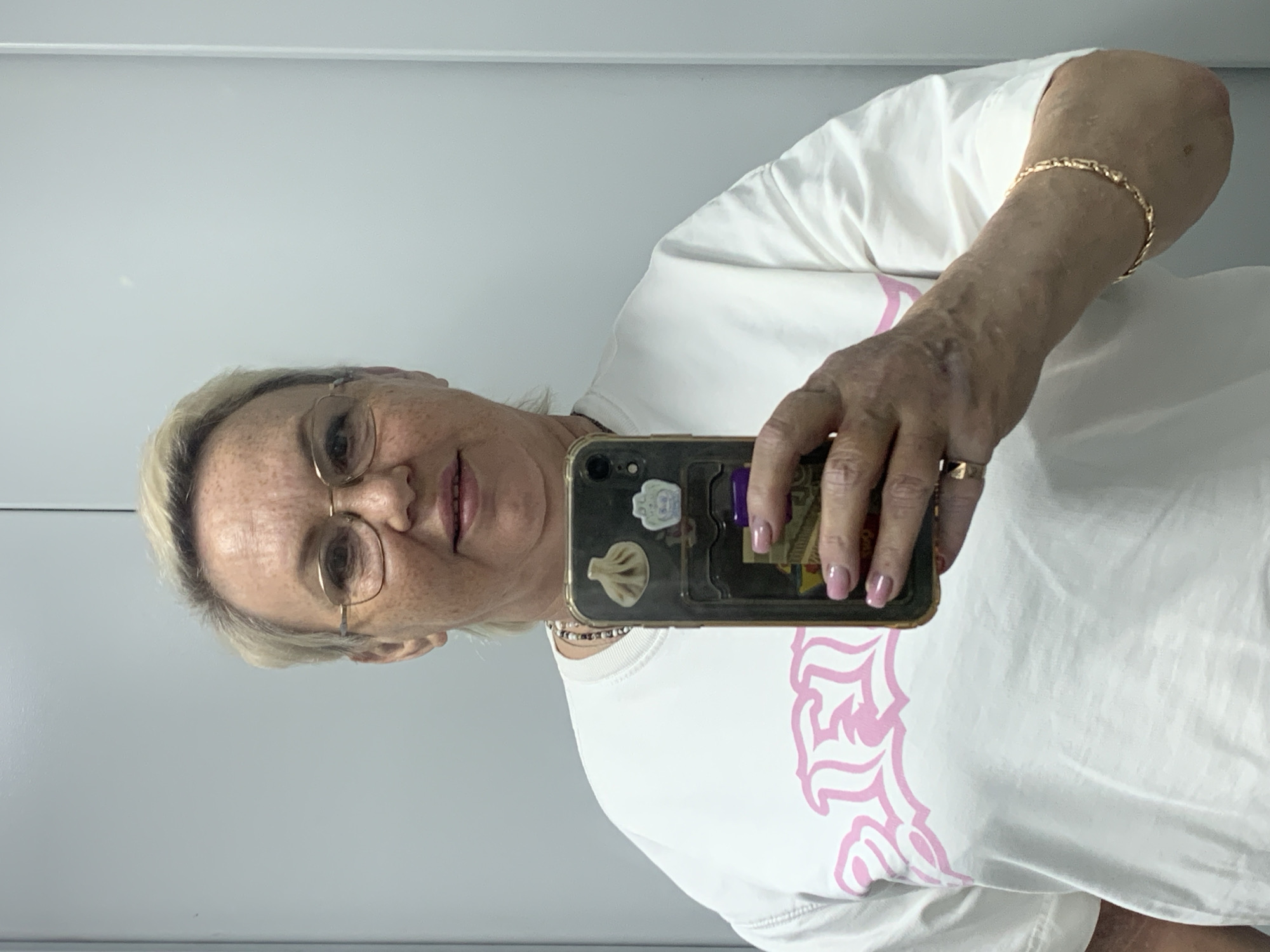 A woman with blonde hair and glasses takes a selfie in a bright, indoor environment, possibly an elevator or a hallway, with plain grey walls. She is wearing a white t-shirt with a pink graphic design on the front. She is also wearing a necklace, a ring on her left hand, and a bracelet on her right wrist. Her phone, which has stickers on its case, is visible in her right hand as she takes the photo. The image does not provide specific clues about its location within the city of Chaikovsky, Russia.