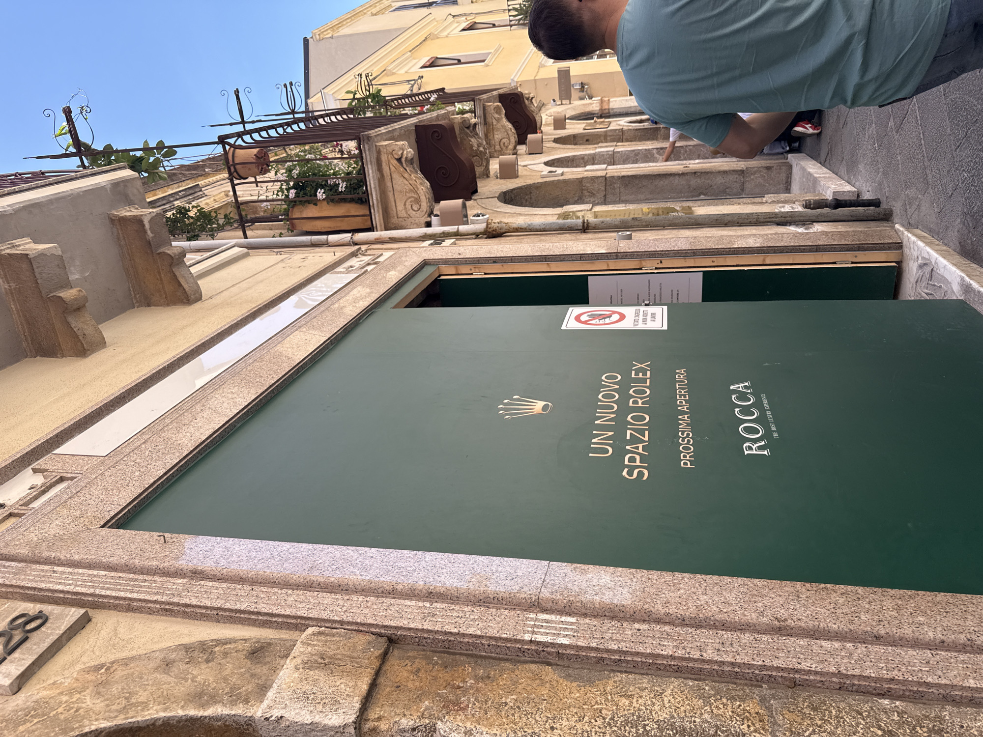 The image shows a partially open storefront in Unknown, Unknown, with a large green door that reads "UN NUOVO SPAZIO ROLEX PROSSIMA APERTURA ROCCA THE BEST LUXURY EXPERIENCE". There is also a sign that reads "VIETATO CONGRESSO A NON ADDET ALAVOR". The storefront is made of tan stone, with a number 28 visible on the left side. Above the storefront are a series of arches and decorative stone carvings, with potted plants visible in some of the arches. A man in a green shirt is crouched down on the sidewalk in front of the store. The sky is blue in the upper left corner of the image.