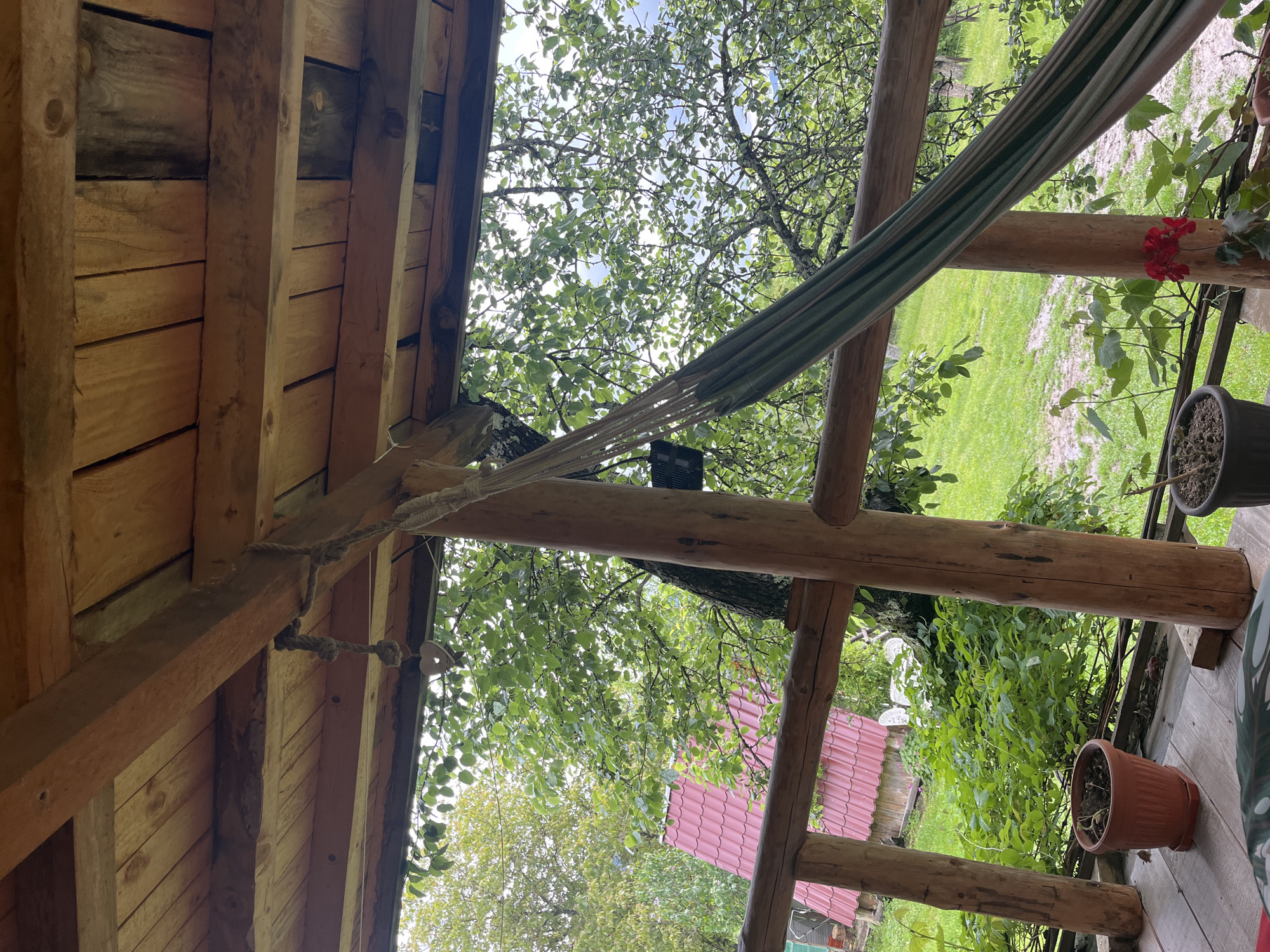 The image depicts a rustic porch setting, potentially in Shopite, Bulgaria, although no clear identifiers confirm the location. A wooden structure forms the porch's frame, with exposed beams and planked roofing. A striped hammock is strung between support posts. Outside the porch, greenery is visible, including trees and foliage, and a building with a red tiled roof. Potted plants are positioned on the porch's edge. The scene appears peaceful and natural.