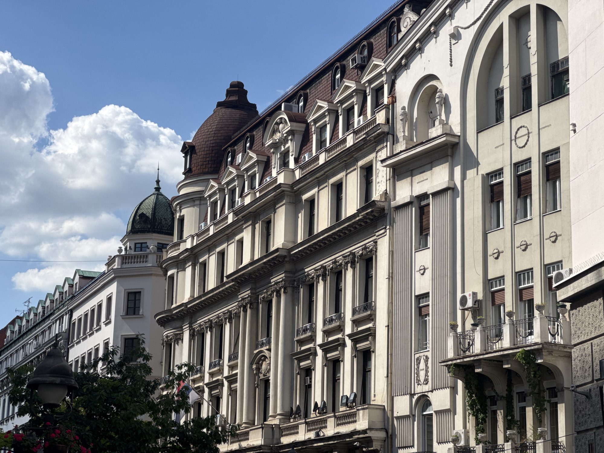 The image displays a bright, sunny day in Belgrade, Serbia, with a clear blue sky and fluffy white clouds. The scene is dominated by the detailed facades of several grand, old buildings, showcasing a mix of architectural styles.
On the left, a lighter-colored building with a green dome and a spire is partially visible, with a row of mansard roofs extending to the left. Next to it, a larger, cream-colored building features a prominent brown mansard roof with dormer windows and decorative gables. This building is characterized by ornate columns, balconies with wrought-iron railings, and numerous windows. Further to the right, another building exhibits a more modernist facade with large arched openings, vertical fluting, and circular decorative elements. This building also has balconies adorned with hanging greenery.
Several air conditioning units are visible on the facades, indicating modern functionality within the historic architecture. The overall impression is one of a bustling urban environment with a rich architectural heritage. The lighting suggests it is daytime, likely late morning or early afternoon, given the bright sunlight and distinct shadows. There are no people clearly visible in the image, and the focus is solely on the architecture and the sky.