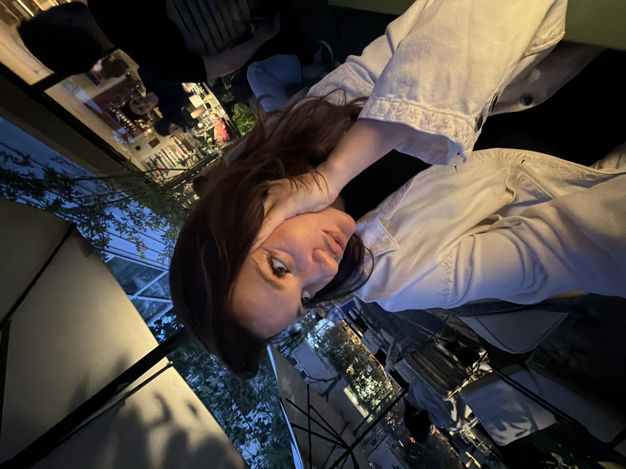 A woman with long auburn hair is captured in a close-up, slightly angled shot. She is wearing a white jacket and a black top. Her hand is raised to her face. Behind her, a blurred outdoor scene includes tables and people at what appears to be a restaurant. An umbrella is partially visible in the foreground. The background suggests a twilight setting with trees and buildings, potentially indicating an urban environment.
The location is not clear enough in this image to determine whether it is in Belgrade, Serbia or not.