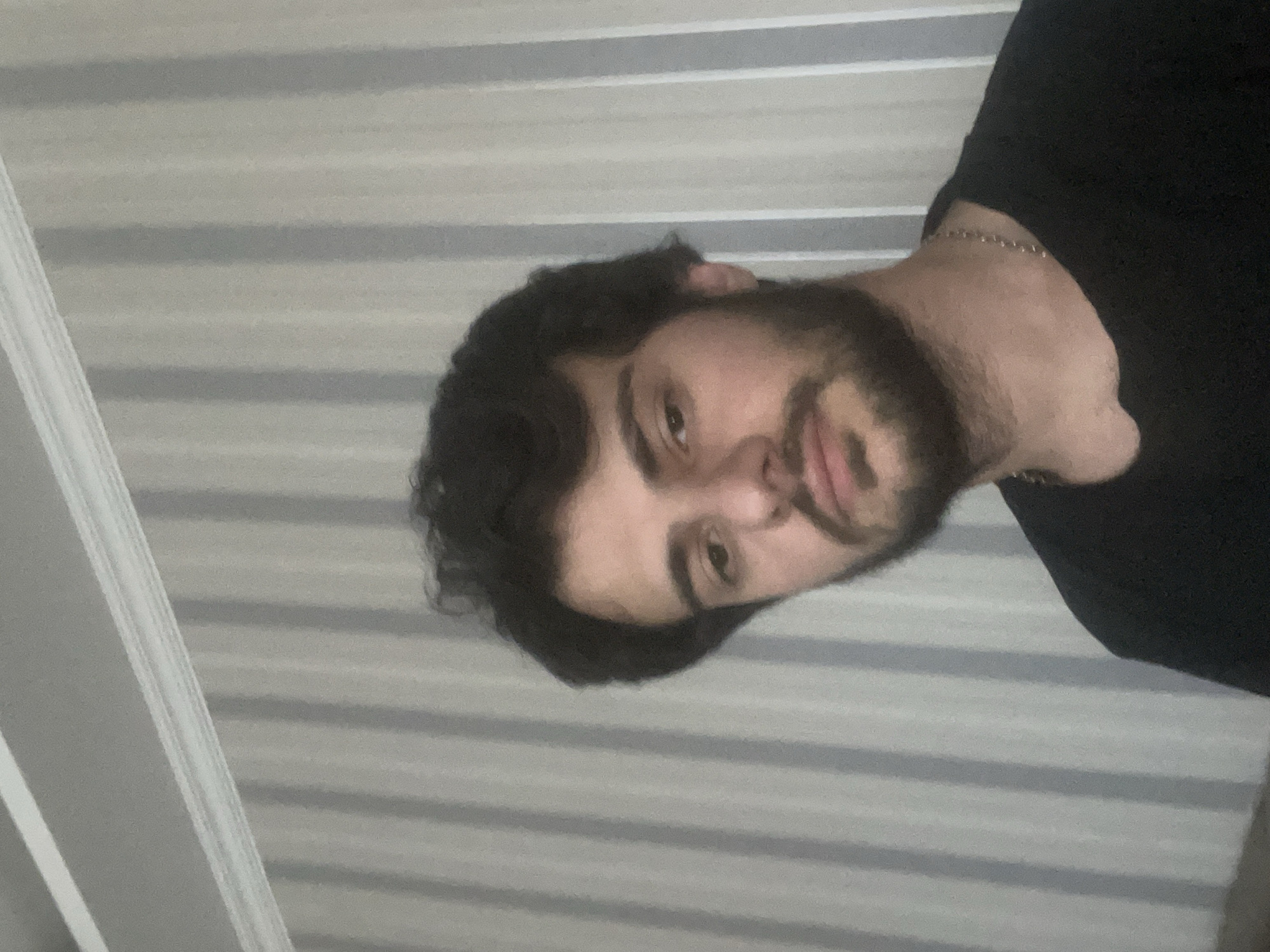 A close-up, head-and-shoulders selfie is taken by a young man with dark, wavy hair and a light beard. He is wearing a black t-shirt and a silver chain necklace. The background consists of a wall with vertical, light gray and white striped wallpaper. Above the wallpaper, a white crown molding is visible, and above that, a white ceiling. The lighting is soft and appears to be from an overhead source. The image does not provide any visual information that would allow for the identification of its location in Tbilisi, Georgia, or any other specific city.