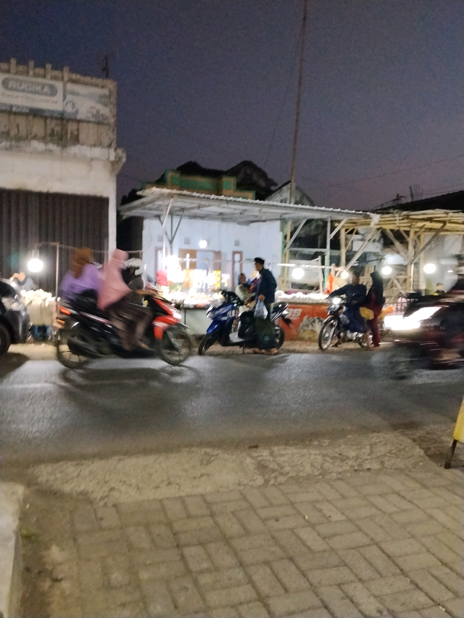 The image depicts an outdoor scene at dusk or night in Panggungan, Indonesia, characterized by low light conditions. In the foreground, a paved street is visible, with a textured, lighter-colored sidewalk to the right. The middle ground features several motorcycles and people. One motorcycle, a red and black scooter, is in motion, carrying two individuals. Another blue motorcycle is parked or moving slowly, with a person on it and another standing nearby. Further back, a market or roadside stall area is illuminated by artificial lights. Structures with white roofs and exposed wooden supports are present, suggesting a temporary or open-air setup. A building with a sign that appears to read "RUGIKA" is to the left, partially obscured by a dark vertical shutter. The sky is dark, with subtle hints of fading light. The overall impression is a lively street scene with ongoing activity in a commercial or public area.