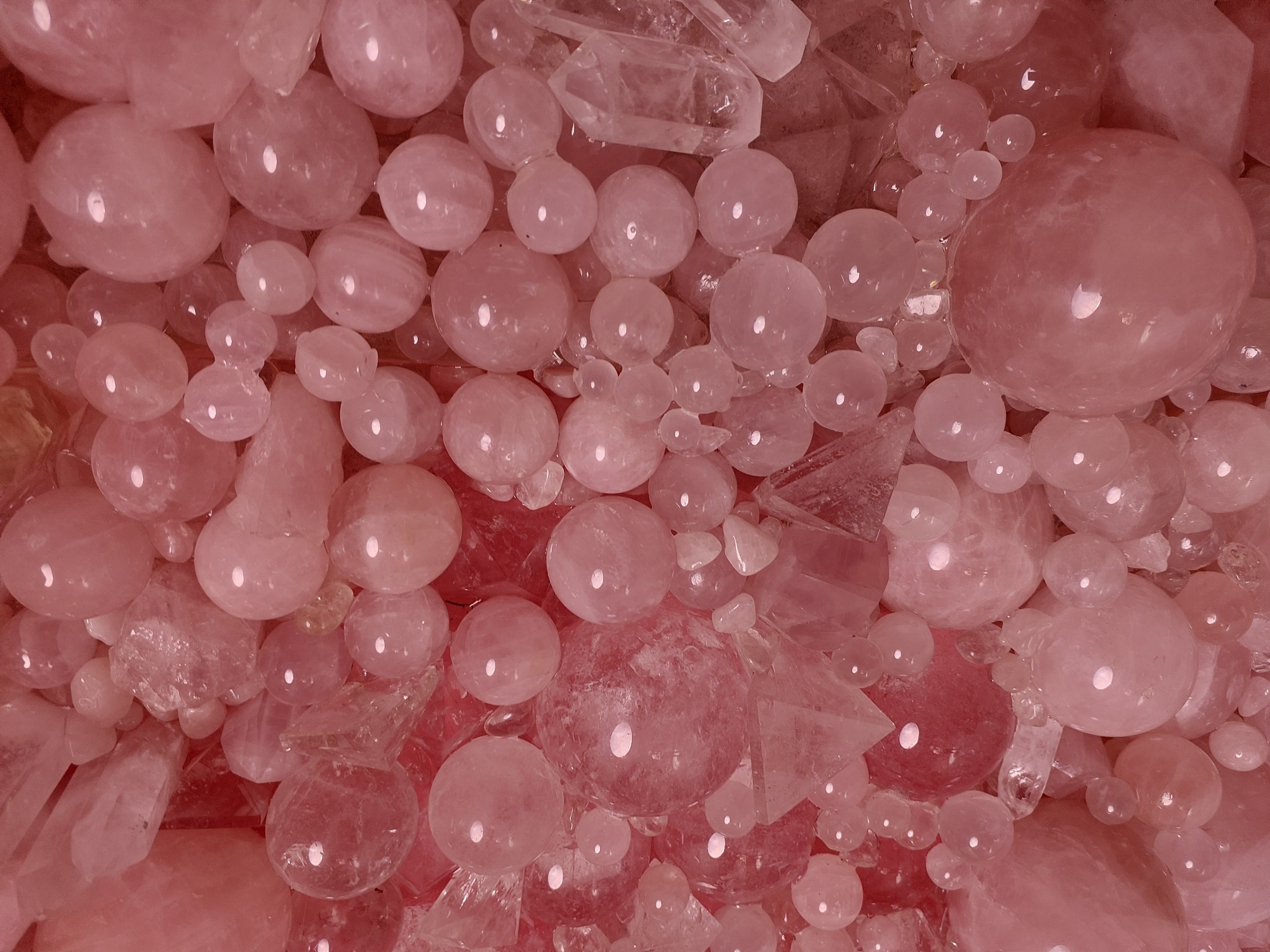 The image displays a dense collection of smooth, spherical rose quartz stones of various sizes, interspersed with rough, crystalline fragments of the same mineral. The stones are a soft, translucent pink, and light reflects off their polished surfaces, creating bright highlights. The composition is a close-up, overhead view, filling the entire frame with the textures and colors of the stones. There is no indication of a specific location in Monaco, Monaco, or any people or actions. The scene is static and purely focuses on the mineralogical arrangement.
