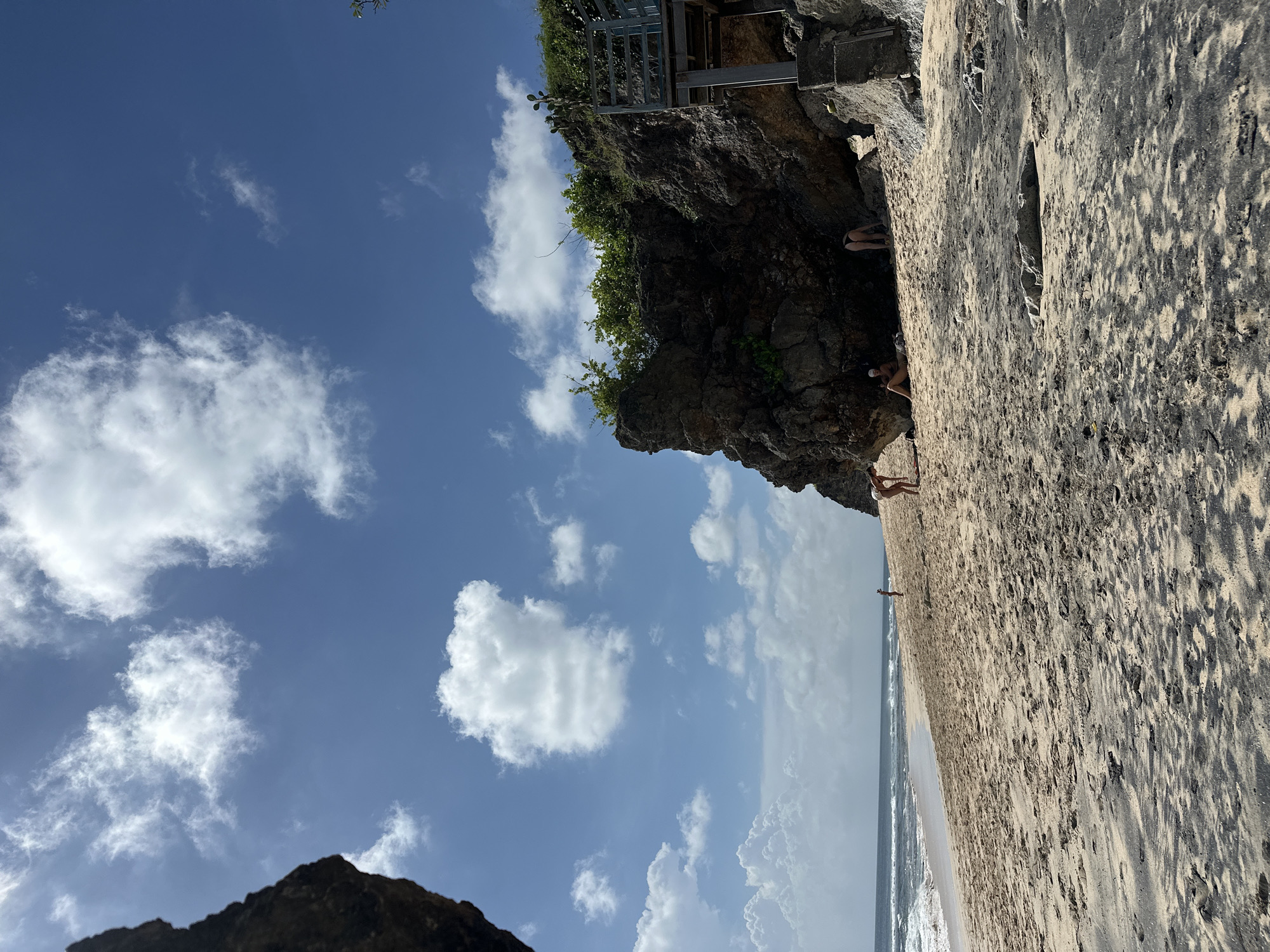 The image depicts a beach scene in Kuta Selatan, Indonesia, notable for its rugged coastline. A rocky cliff topped with vegetation and a metal structure borders a sandy beach where several people are present. The sky is predominantly blue with scattered white clouds. The beach shows varying shades of sand, and the ocean's edge is visible.