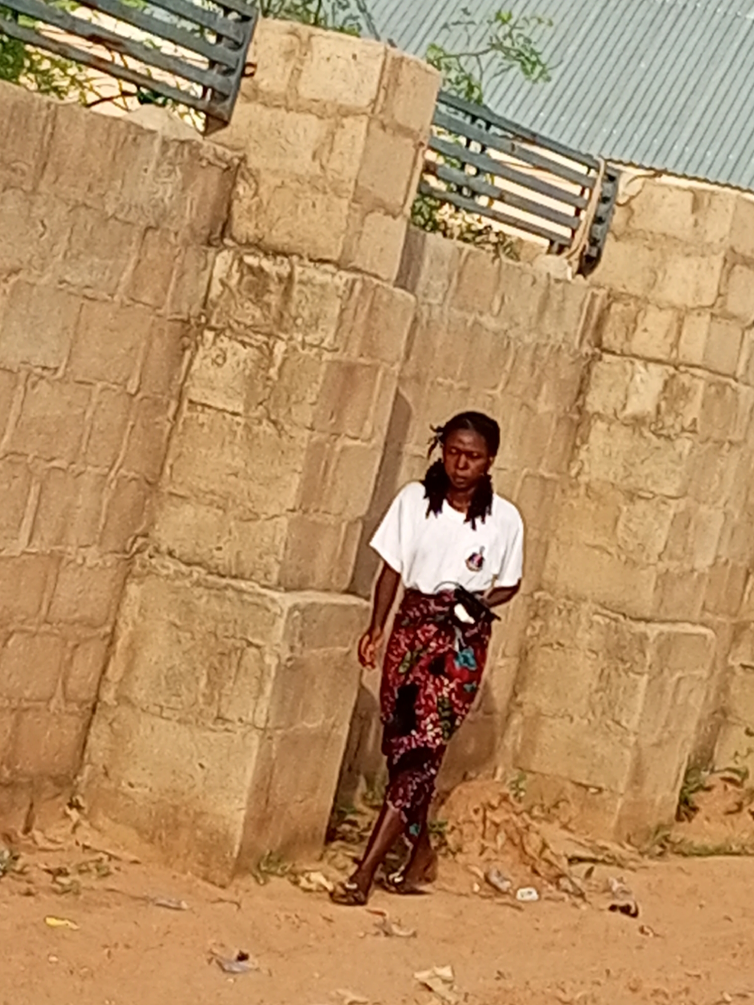 A woman with dark skin and braided hair walks in front of a rough stone wall. She is wearing a white t-shirt and a patterned skirt. The ground is dirt and sand, with some debris scattered around. The wall is made of large, unevenly cut stones. A metal fence with horizontal bars is visible above the wall, with some greenery behind it. The scene appears to be outdoors in a warm climate. The location is identified as Tempure, Nigeria.