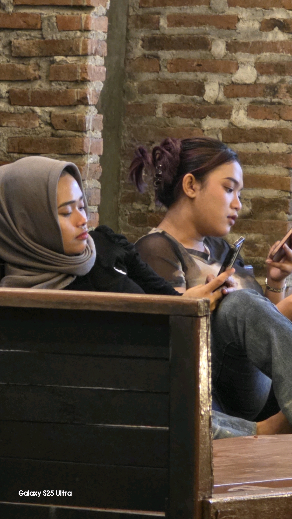 The image shows two young women seated next to each other, possibly in a cafe or restaurant in Juwana, Indonesia. The woman on the left wears a grey hijab and a black top. She is looking down with her eyes closed. The woman on the right has her hair tied up in a bun and wears a sheer patterned top with jeans. She is looking at her phone, which she holds in her right hand. Her left hand is also holding a phone. The background consists of a brick wall. The bottom left of the image displays the text "Galaxy S25 Ultra".