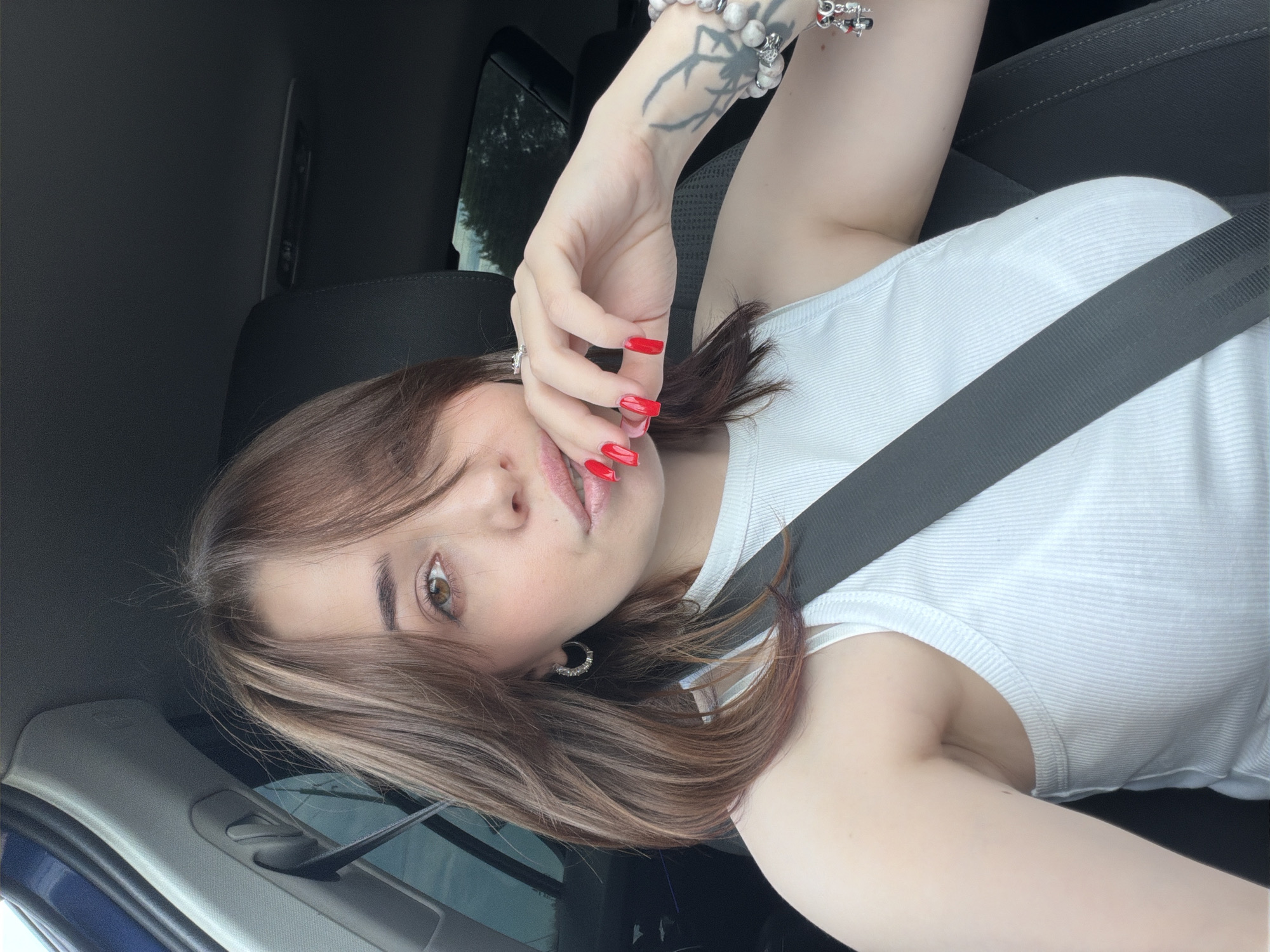 This media file shows a close-up, selfie-style shot of a young woman sitting in a car. She is wearing a white ribbed tank top and a black seatbelt crosses her chest. Her right hand is raised and resting on her face, with long, red-painted fingernails. She has brown hair with lighter highlights, styled to frame her face. Her makeup includes eyeliner and lip gloss. She is wearing a silver hoop earring in her visible ear and a bracelet with charms on her wrist, which also has a tattoo on it.
The lighting suggests it is daytime, and the view through the car window shows greenery, indicating an outdoor setting. The image is taken from a low angle, looking up towards the woman's face. The overall emotion conveyed is casual and relaxed. No visible text is present in the image.