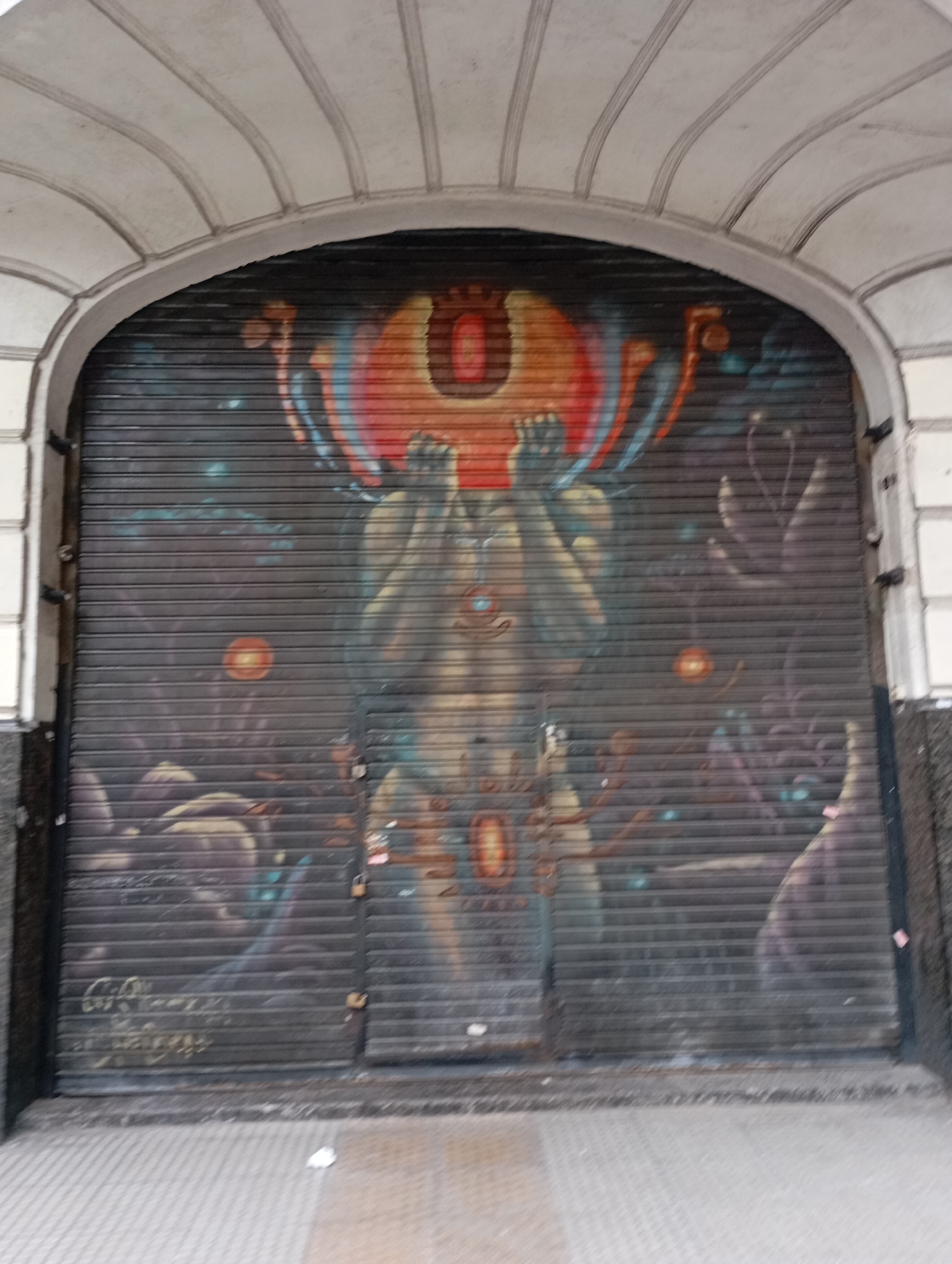 The image shows a large, rolling metal security gate that has been painted with a mural. The mural depicts a muscular, semi-nude humanoid figure with its hands clasped over its face. The figure's head is replaced by a large, circular, orange and red object that resembles an eye or a sun, with blue and yellow accents. Surrounding the central figure are darker, shadowy forms that appear to be other figures or monstrous creatures. The background is a dark, star-like pattern. The gate is situated under a decorative, arched architectural feature, suggesting it is the entrance to a building. The overall scene is dark and somewhat ominous. The specific location is on a building in the Monserrat neighborhood of Buenos Aires, Argentina, specifically on Avenida Corrientes, a well-known street famous for its theaters and nightlife.