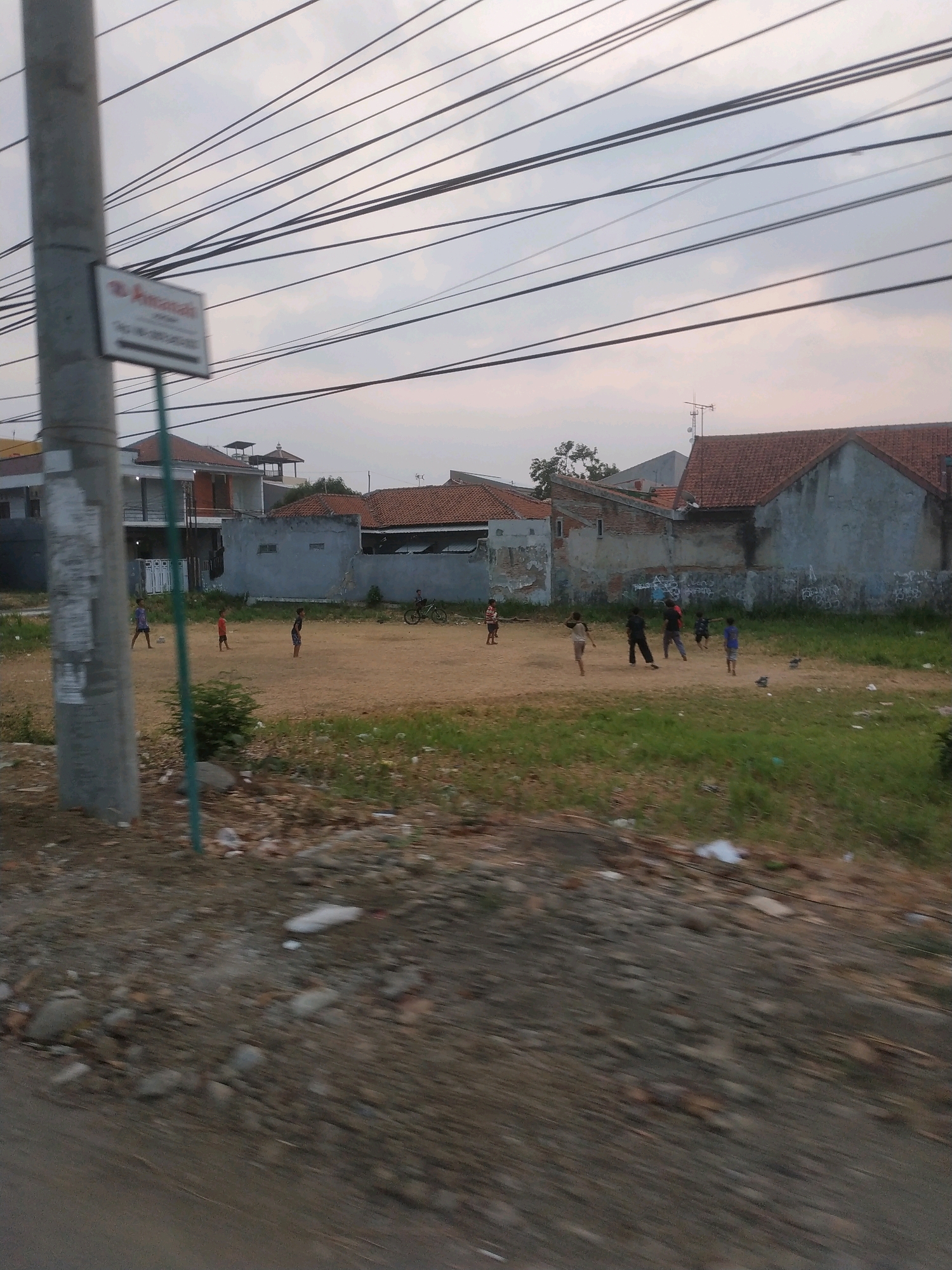 The image depicts an outdoor scene with several children playing soccer on a dirt field. In the background, there are residential buildings with tile roofs, characteristic of urban or suburban areas in Tegal, Indonesia. Utility poles and numerous overhead wires are visible, suggesting the presence of electricity and telecommunications infrastructure. The ground is a mixture of dirt and sparse grass, with some scattered debris. The sky is overcast with a soft, diffused light, indicating it could be dawn or dusk. The overall impression is of a casual, everyday scene in a residential neighborhood.