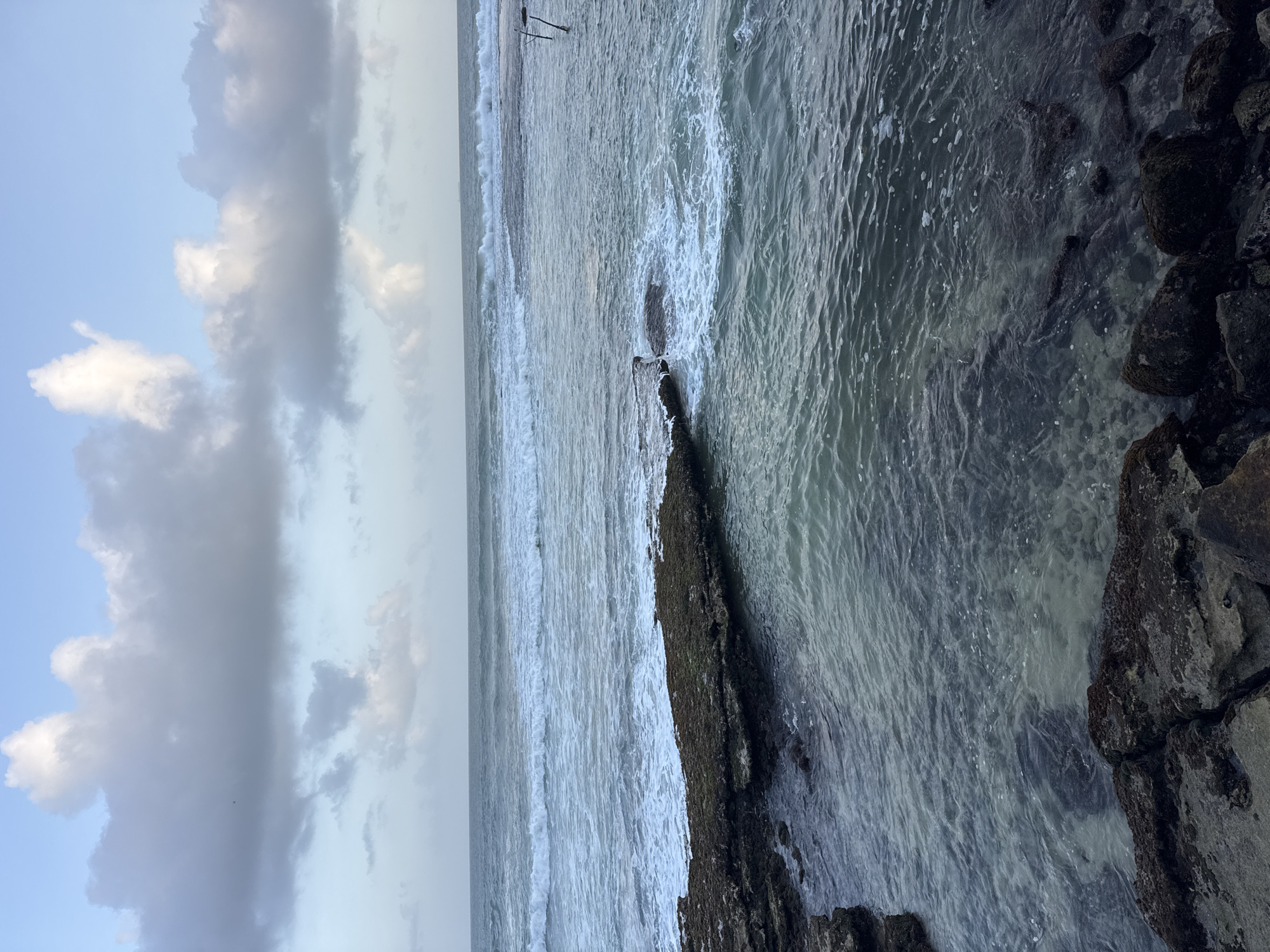 The image depicts a rocky coastline on a cloudy day, likely in Goviyapana, Sri Lanka. The foreground shows dark, weathered rocks, some submerged in shallow, clear water, revealing the sandy bottom. Further out, the ocean stretches to the horizon, with moderate waves breaking against a rocky outcrop and dissipating into white foam. In the distance, the sea appears rougher, with more pronounced waves. The sky is filled with a mix of light and dark grey clouds, with patches of blue sky visible. Two thin, dark vertical poles, possibly remnants of a fishing structure, are present in the water on the right side of the frame. The overall mood is serene and natural, with no people or animals visible.