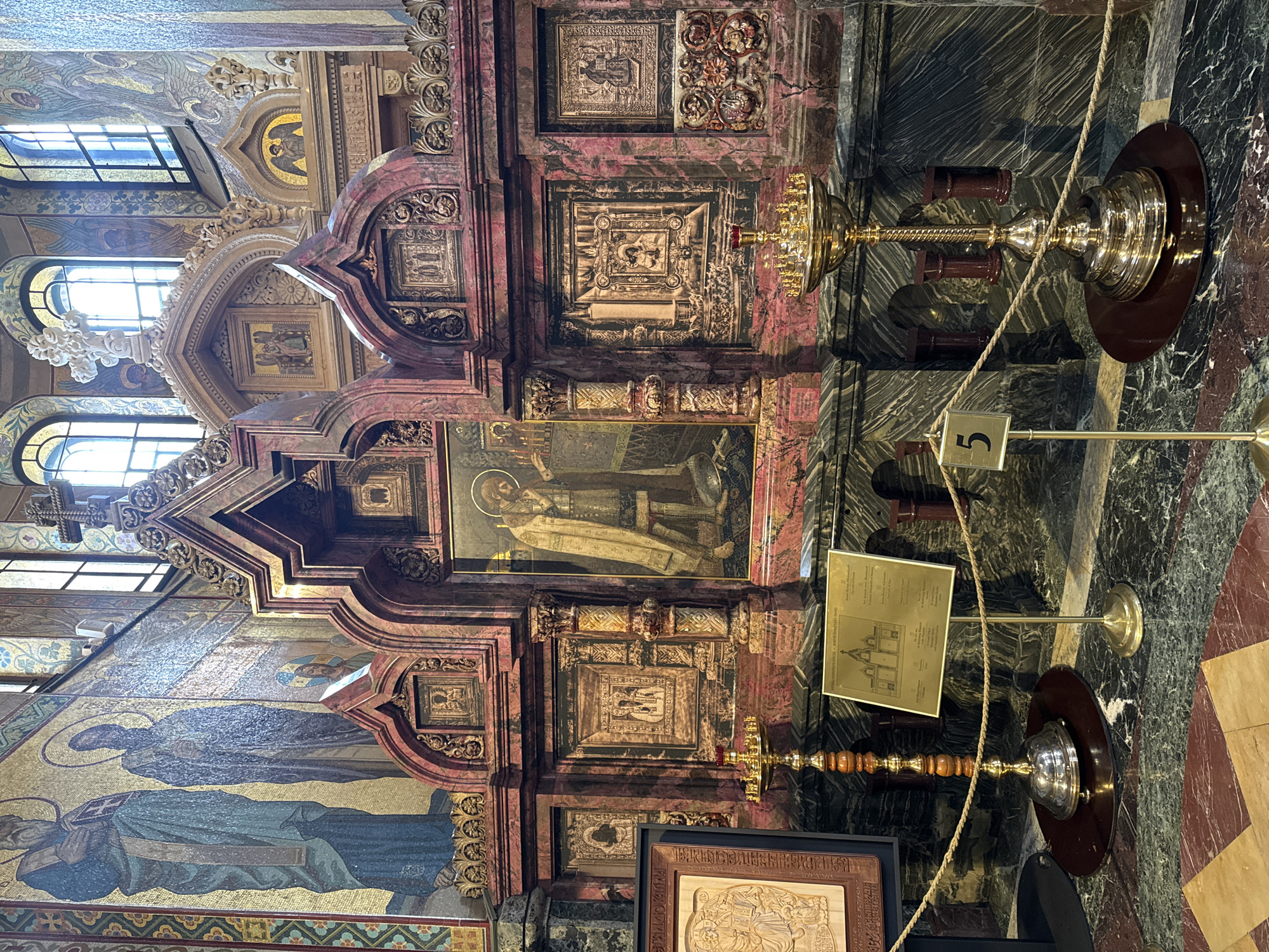 The image captures an interior shot of the Church of the Savior on Spilled Blood, located in Saint Petersburg, Russia. The viewpoint is focused on a section of an ornate, mosaic-filled wall featuring religious figures. Architectural details include archways, framed panels, and decorative carvings. A vertical display stand with a number "5" is visible, as well as candle stands. A rope barrier is present to prevent visitors from getting too close to the altar. The interior is heavily decorated with gold, mosaics, and colorful stones, which reflect the church's distinctive Russian Revival style.