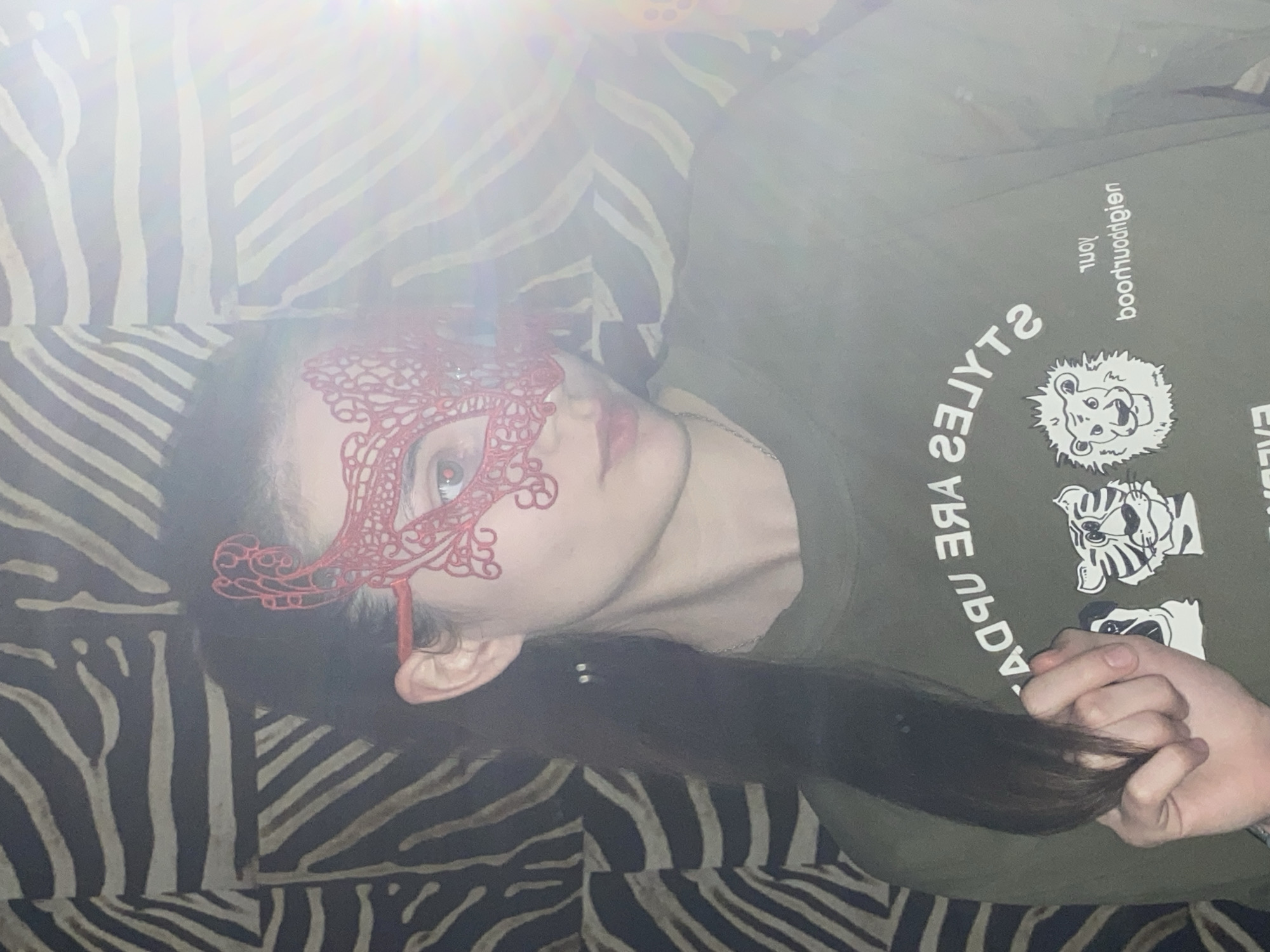 A medium-close shot captures a person lying down, wearing a decorative red masquerade mask that covers the upper part of their face, predominantly their left eye. The visible right eye has a light-colored iris. The person's dark, long hair extends down their back, and they are holding a section of it with their right hand.
They are wearing a dark olive-green t-shirt featuring white graphics and text. The text "STYLES ARE" is clearly visible. Below this, inverted text reads "ruoY" (which is "Your" reversed) and another mirrored word, "boohuoqgiej," which is hard to decipher but appears to be a stylized brand name or phrase. Two animal head illustrations are also present on the shirt: a lion's head and a tiger's head.
The background consists of a prominent black and white zebra-print pattern, likely a couch or bedding, providing a striking contrast. A bright lens flare is visible in the upper left portion of the image, contributing to an overall soft, diffused lighting effect. The image is oriented with the person lying horizontally, as if rotated 90 degrees clockwise from a typical portrait orientation. The setting is indoors, with no specific time of day or weather discernible. The location context is Votkinsk, Russia.