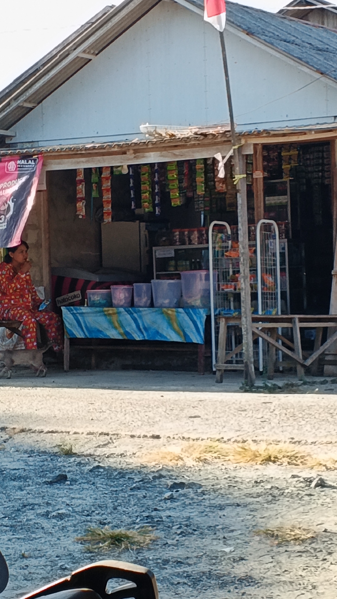 In Kota Pagatan, Indonesia, a woman in a red and white patterned outfit sits on a bench outside a small, open-fronted shop. She appears to be eating or holding something to her mouth. Next to her, on the ground, is a small, light-colored dog. The shop is filled with various packaged goods hanging from the ceiling and displayed on shelves. In front of the shop, a table is covered with a blue and yellow striped cloth, holding several transparent plastic containers of varying sizes. A metal shelving unit with items on display stands to the right of the shop entrance. A flagpole with the Indonesian flag, red and white, is visible to the right of the shop. The ground in front of the shop is a mix of gravel and dirt, with patches of dry grass. A portion of what appears to be a motorcycle is visible in the foreground. The scene is illuminated by sunlight, casting shadows on the ground.