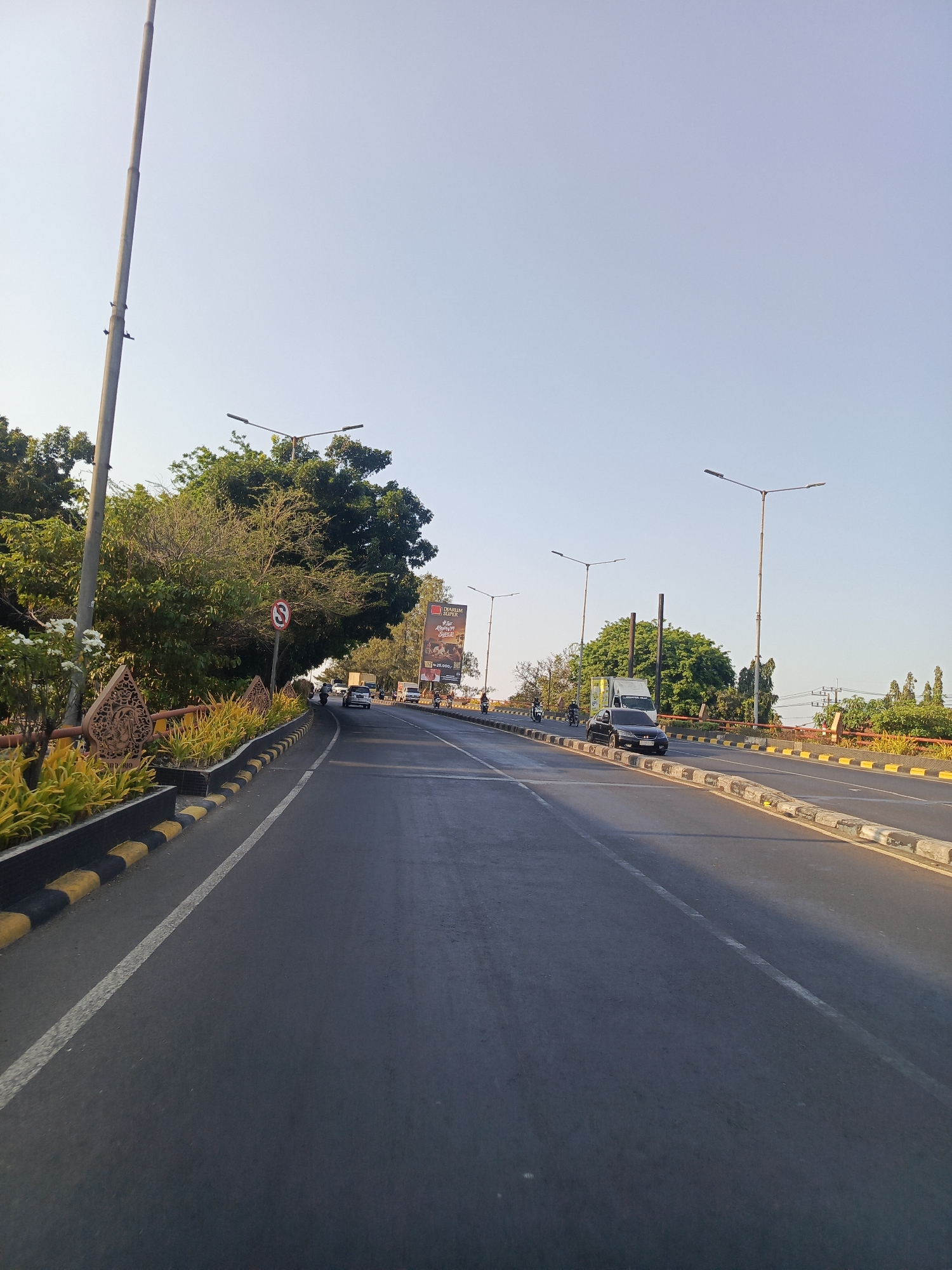 This is a medium-shot, eye-level, outdoor, daytime, wide-angle photograph of a road in Sidokerto, Indonesia. The road is paved and appears to be a highway or major thoroughfare, with multiple lanes and a clear sky overhead.
On the left side of the road, there is a sidewalk with yellow and black striped borders, lined with decorative planters containing yellow and green foliage and a few white flowers. Beyond the sidewalk, there are trees and a tall, metal pole that could be a flagpole or lamppost. A red circular sign with a white 'S' symbol is visible on a pole.
On the right side of the road, there are more planters and trees. A billboard with text and images is visible in the background. Several vehicles are on the road, including cars and motorcycles. A white truck is in the right lane, and a black car is in the left lane. The road curves slightly to the right in the distance. The overall scene depicts a well-maintained road in an urban or semi-urban area with a clear, bright sky and visible streetlights.