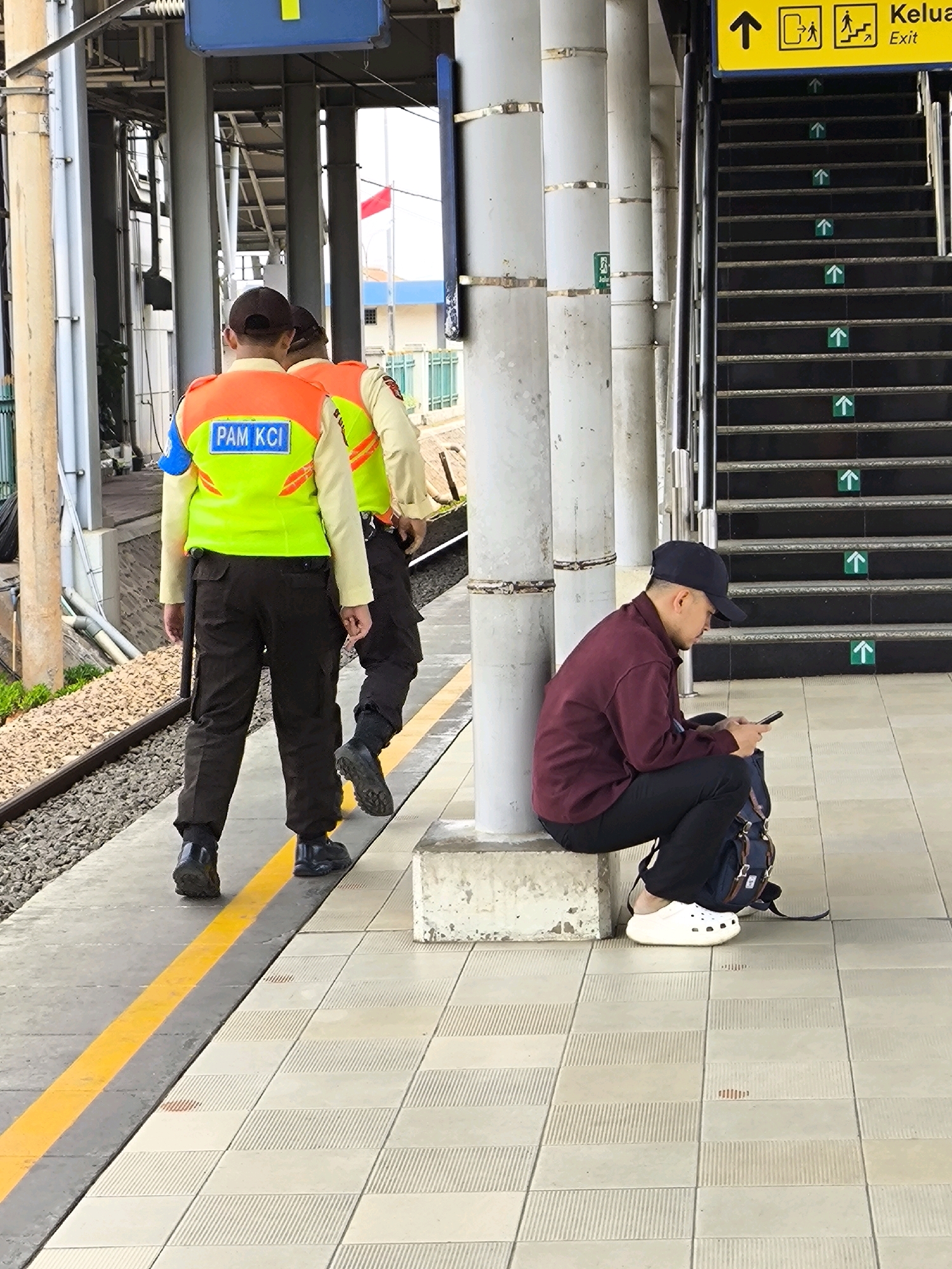 The image depicts a scene at a train station in Mekarsari, Indonesia. In the foreground, two individuals in uniform are walking away from the camera on a tiled platform. The person on the left wears a bright green safety vest with "PAM KCII" printed on the back in blue and orange lettering, over a light-colored long-sleeved shirt and dark trousers. They have a dark baton clipped to their belt. The second person, partially obscured, also wears a similar vest over a light-colored shirt and dark trousers.
To the right, a young man wearing a dark baseball cap, a maroon long-sleeved shirt, and black pants sits on a concrete ledge, engrossed in his smartphone. He is wearing white Crocs-style footwear and has a dark backpack next to him. Behind the seated man, a wide staircase with black treads and metal railings ascends. Small green arrow stickers indicating upward direction are placed on each step. An overhead yellow sign with black text and icons, including an upward arrow and figures representing people walking and climbing stairs, is visible above the staircase, with "Exit" written in English. The environment appears to be an outdoor covered area of a train station, with concrete pillars and elevated tracks visible in the background. The ground on the platform consists of light-colored tiles, with a yellow line marking a boundary near the train tracks, which are covered in ballast. The overall lighting suggests an overcast day.