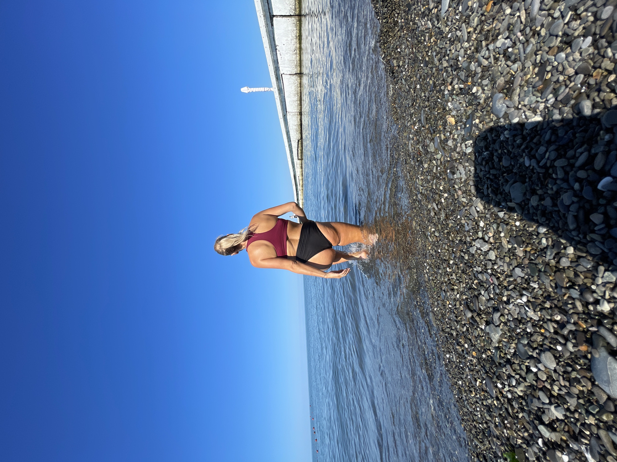 A woman walks into the sea on a pebble beach. She is wearing a maroon bikini top and black bikini bottoms. The sky is clear blue. A concrete pier with a white structure on it is visible in the background. The sea is calm with small waves. The beach is covered in dark grey and brown pebbles.