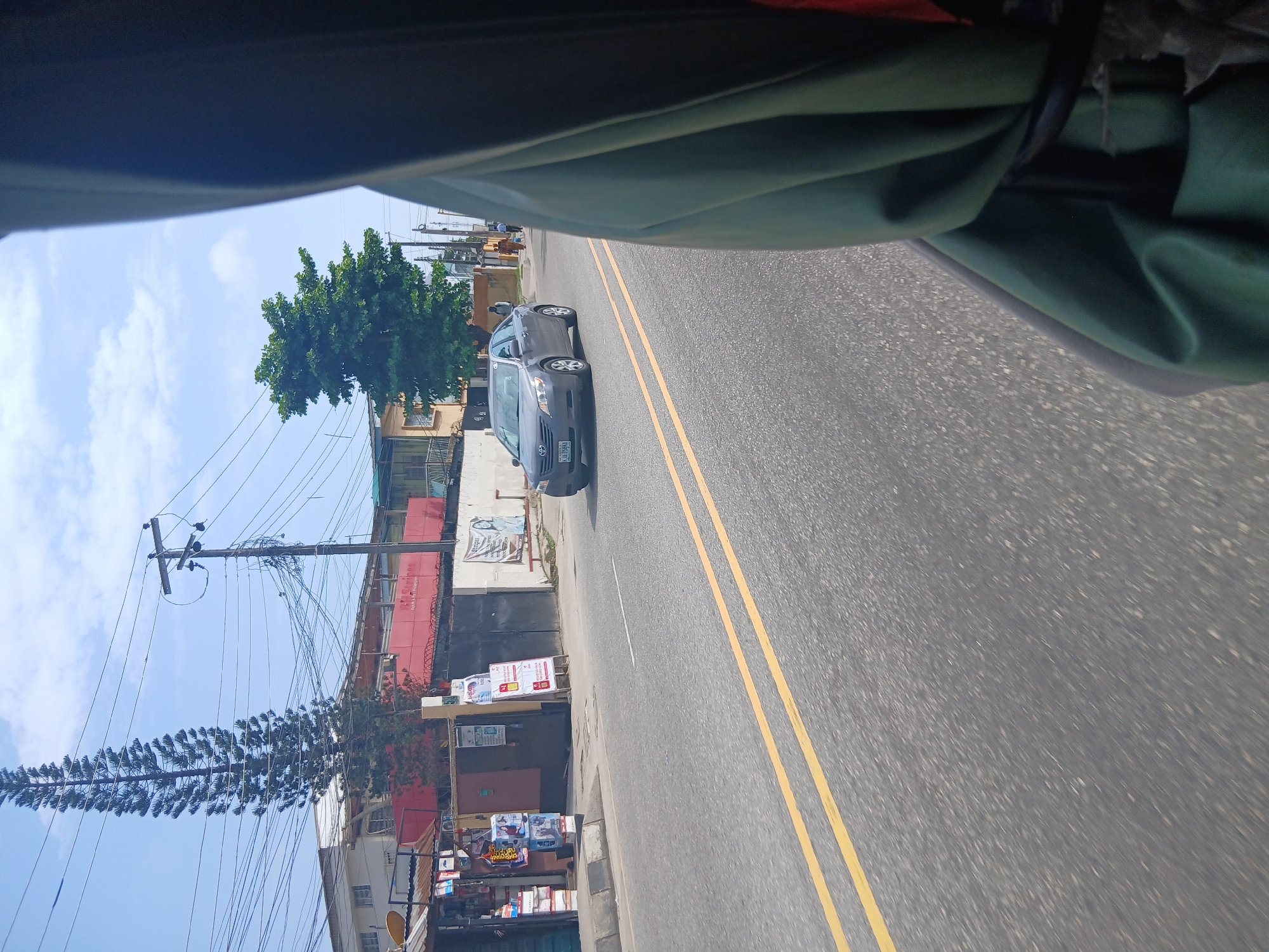 A street scene in Animashawun, Nigeria, is captured from inside a moving vehicle, with a portion of the vehicle's interior and a green tarp obscuring the right side of the frame. The asphalt road features double yellow lines indicating the lane dividers. A grey sedan, license plate KAV 596A, is traveling in the oncoming lane, its headlights on. Several pedestrians are visible on the sidewalk in the distance. Buildings line the street, including one with a red facade and a tall, slender evergreen tree in front of it. Another building has a lush green tree with a wider canopy. Utility poles and a dense network of overhead wires dominate the sky. Signs and posters are displayed on some of the buildings and on freestanding stands along the sidewalk. The sky is partly cloudy.