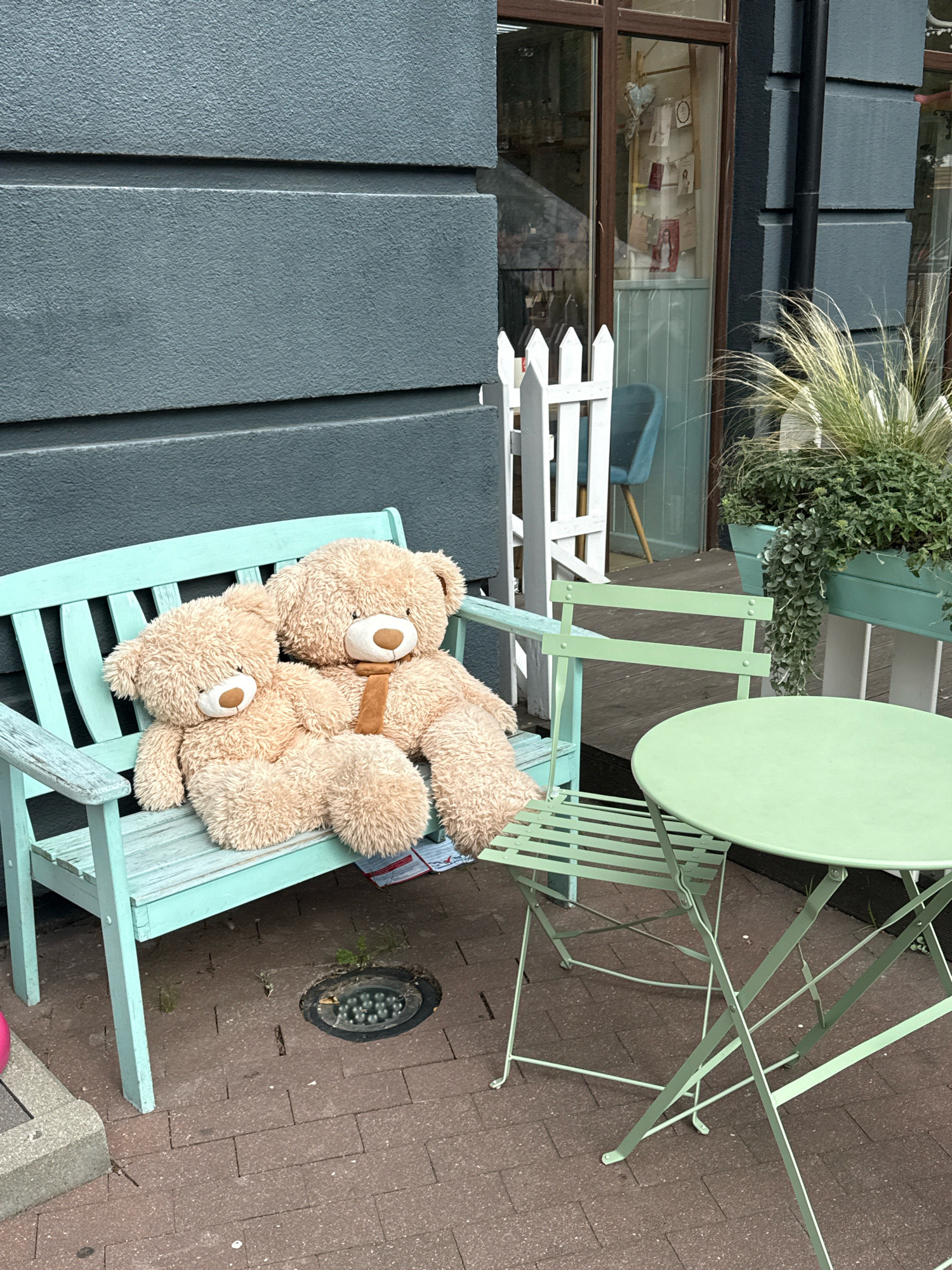 Two teddy bears are sitting on a light blue wooden bench outside a building. The teddy bears are light brown and fluffy. The bear on the right has a brown tie around its neck. To the right of the bench is a light green metal table and chair set. The table is round and the chairs are slatted. In the background, there is a white picket fence, a window of a building, and plants in a green planter. The scene appears to be an outdoor seating area in a city. The image does not provide specific information to determine the city of Svetlogorsk, Russia.