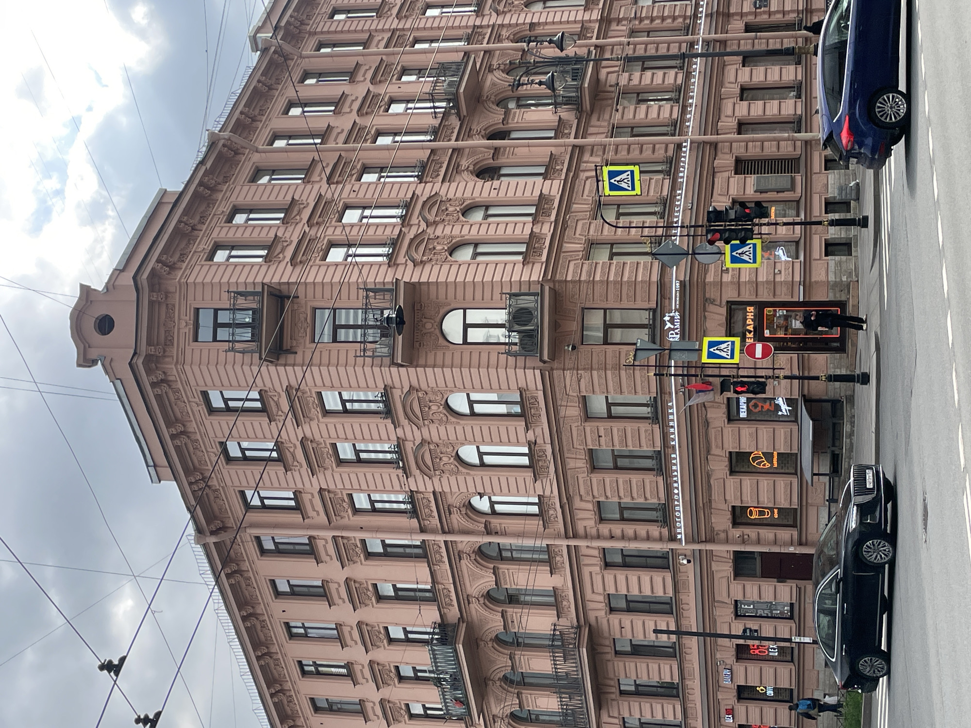 The image depicts a street scene in Saint Petersburg, Russia. A tall, ornately decorated pink building dominates the left side of the frame. The building has numerous windows and detailed architectural features such as balconies and decorative moldings. Streetlights with pedestrian crossing signs are visible along the sidewalk. Two cars are parked on the right side of the street, one blue and the other dark. There is commercial signage, including what appears to be bakery and cafe, on the ground floor of the building. Electrical wires crisscross the sky above. The sky is overcast with grey clouds. A person is walking on the sidewalk near the commercial signs.