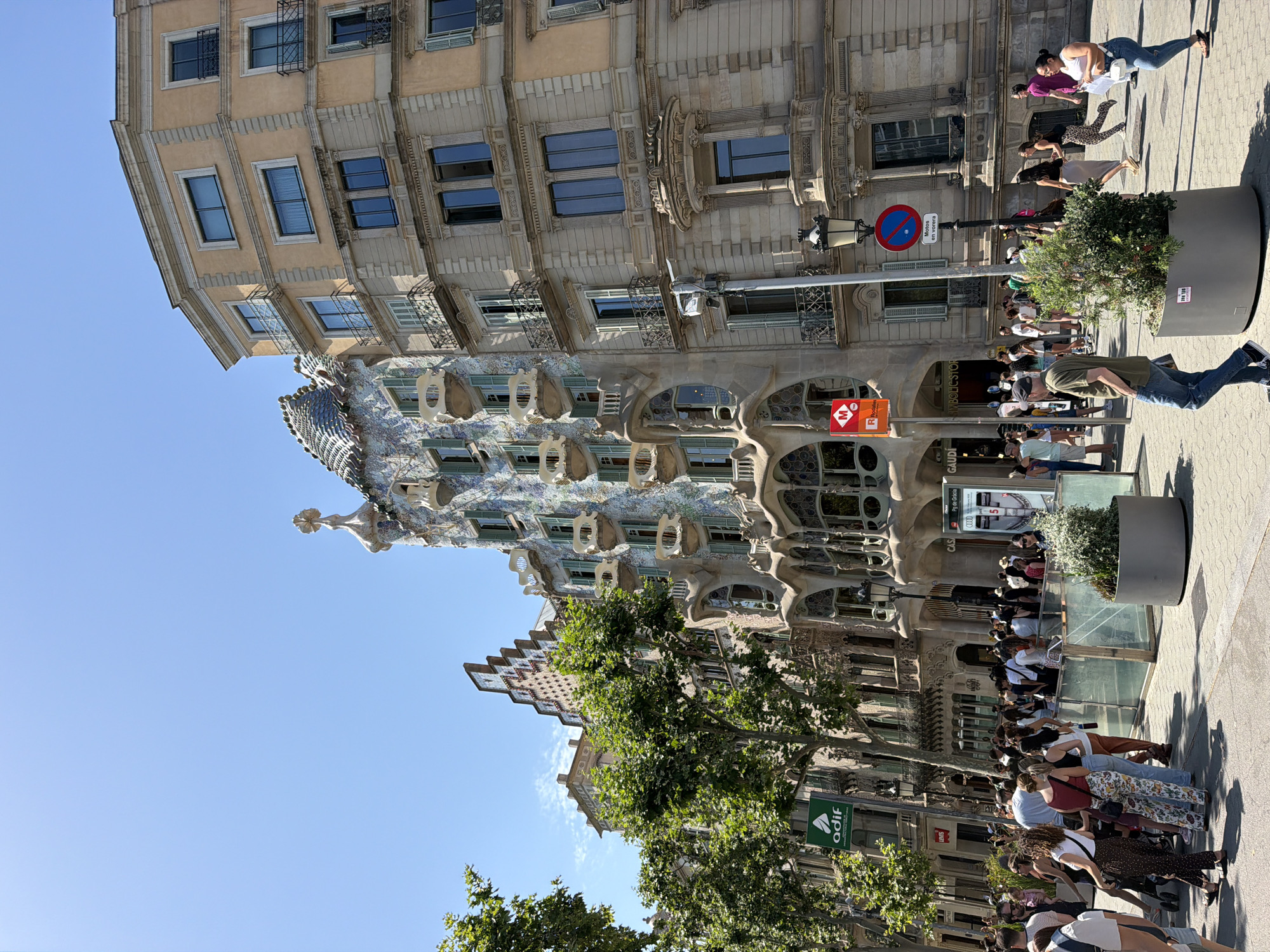 The image depicts the exterior of Casa Batlló, a renowned architectural work by Antoni Gaudí, located on Passeig de Gràcia in Barcelona, Spain. The building's distinctive facade, characterized by its colorful mosaic tiles and organic, flowing shapes, is prominently featured. Adjacent to Casa Batlló is a more traditional, multi-story building with light-colored stone and balconies. A crowd of tourists is visible in the foreground, gathered on the sidewalk, suggesting a popular tourist destination. Several trees with lush green foliage line the street. Various street furniture, including lampposts, signage (one displaying "GAUDI"), and planters, are also present. The sky is clear and blue, indicating a sunny day. The scene captures a vibrant urban environment with a focus on the unique architecture and the flow of pedestrian traffic.