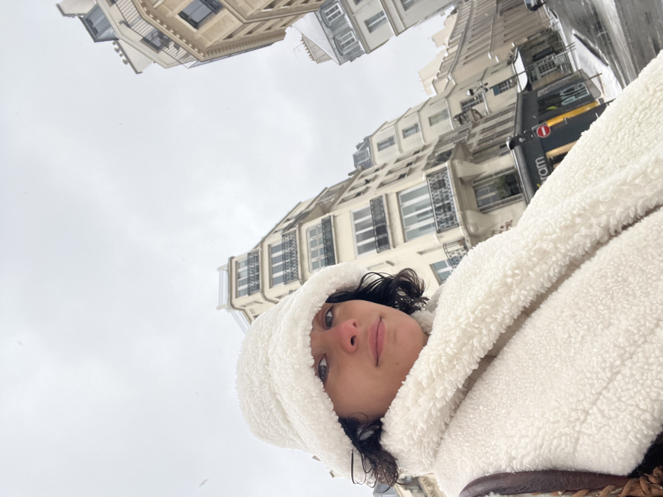 The image features a female subject in the foreground, captured in a low-angle perspective against an urban Parisian backdrop during snowy conditions. The woman has short, dark, curly hair and looks directly at the camera with a neutral expression. She is wearing a white, fluffy bucket hat and a matching white, textured scarf or jacket. Her nose appears slightly reddened.
The background reveals a street scene in Paris, France, characterized by multi-story Haussmannian buildings. These structures display light-colored stone facades, prominent black wrought-iron balconies, and numerous windows. Snow partially covers the rooftops, balconies, and window ledges of these buildings. The sky is uniformly overcast and grey, with visible snow flurries. The street and sidewalks are covered with a thin layer of slushy snow. On the right side, at street level, a portion of a commercial establishment's storefront is visible, showing partial text "ram" and "Pic," along with a red circular "no entry" street sign mounted on a pole. The overall scene depicts a cold, winter day in the city.
