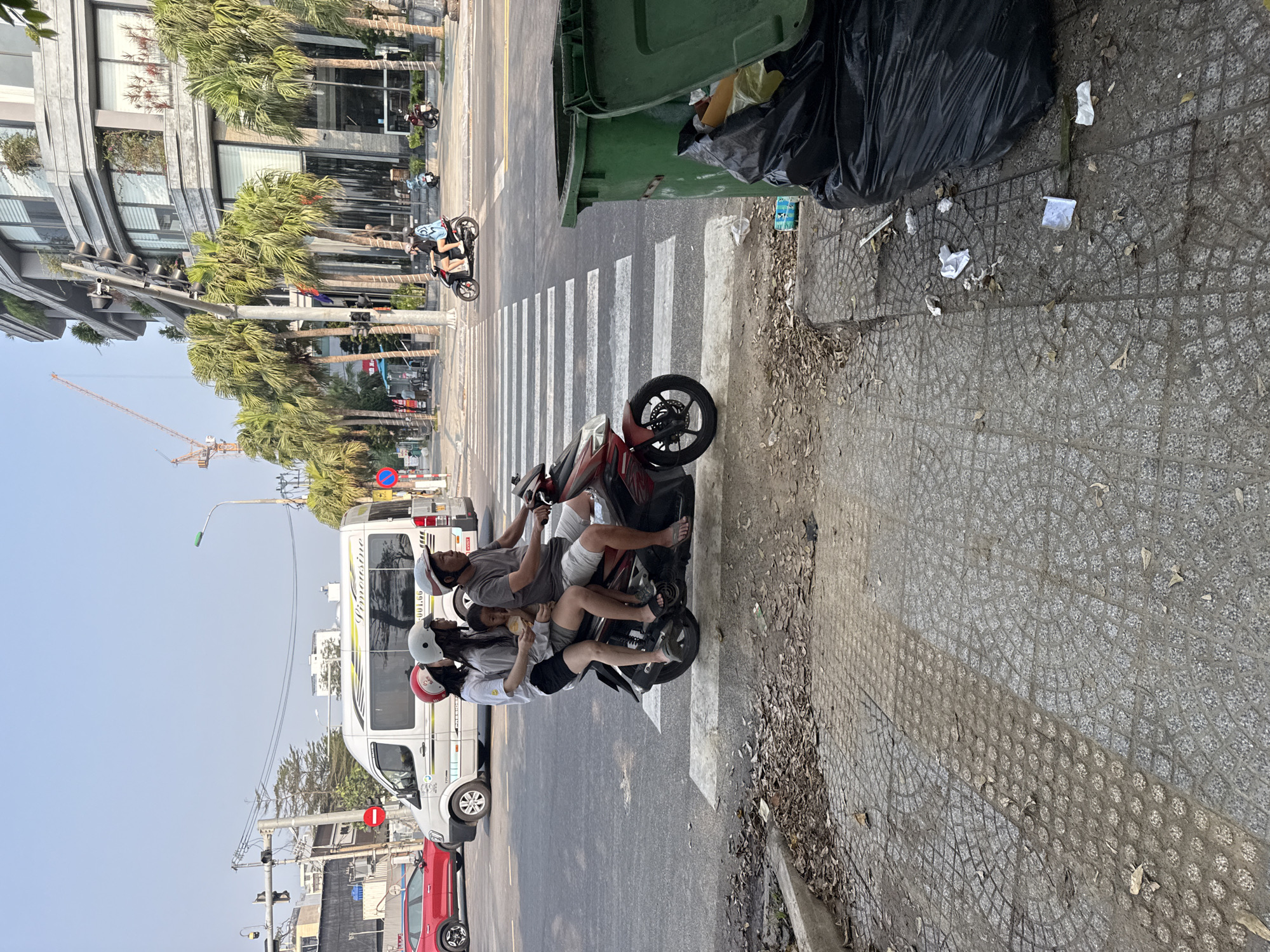 A family of four rides a red scooter across a crosswalk in Đà Nẵng, Vietnam. The father drives, wearing a grey t-shirt and no shoes. The mother is behind him, wearing a white shirt, black shorts, and a red helmet. There are two children on the bike; one appears to be eating a snack. The scooter is on a crosswalk. A green garbage bin filled with trash sits on the sidewalk near the scooter. Litter is scattered on the sidewalk. A white van and a red car are parked on the side of the road. In the background, there are buildings and palm trees.