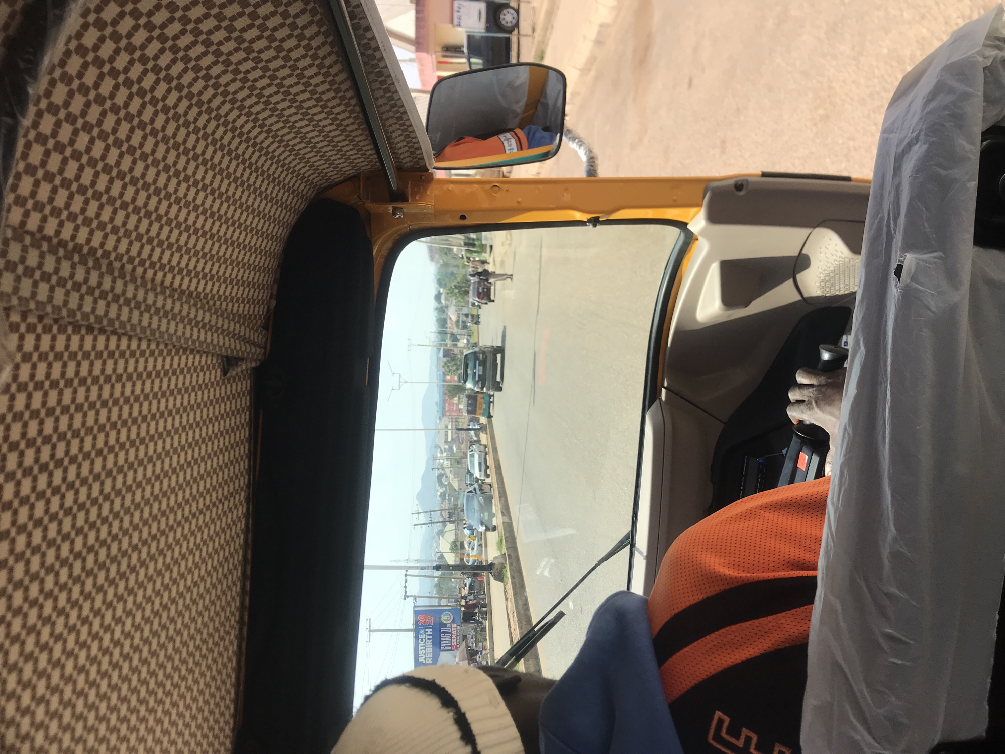 The image shows the interior of a yellow auto-rickshaw driving down a street in Jos, Nigeria. The ceiling of the auto-rickshaw is lined with a checkered fabric pattern. The driver, wearing a blue hoodie and an orange and black striped shirt, is visible from the back, with his hand on the steering wheel. A white knitted cap is on his head. Outside the vehicle, the street is paved and lined with sidewalks. Vehicles, including other auto-rickshaws, cars, and a truck, are driving on the road. Buildings and shops are visible on either side of the street, with some displaying advertisements. A billboard with "JUSTICE & 20 REBIRTH 20" is prominent on the left side. In the background, hills and mountains can be seen. The sky is clear and bright.