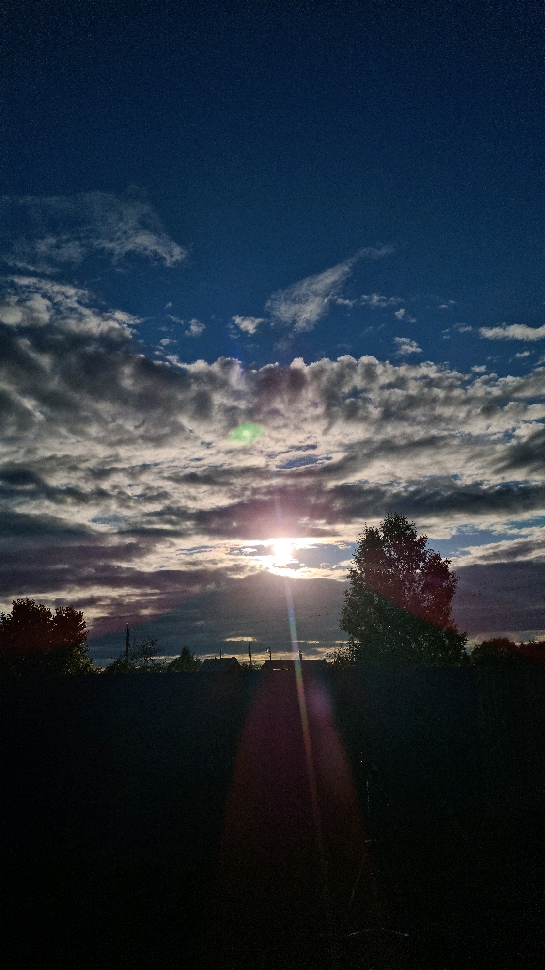 The image depicts a dark foreground with a silhouetted fence, possibly in the city of Vysokoe, Russia. Beyond the fence, the landscape transitions into a dimly lit scene with trees and buildings. The sky is predominantly dark blue, with a dramatic display of clouds illuminated by a bright sun peeking through. The sun is positioned in the middle of the frame, casting rays and lens flares across the image, suggesting it is either sunrise or sunset. The overall impression is one of a rural or semi-rural environment during twilight.