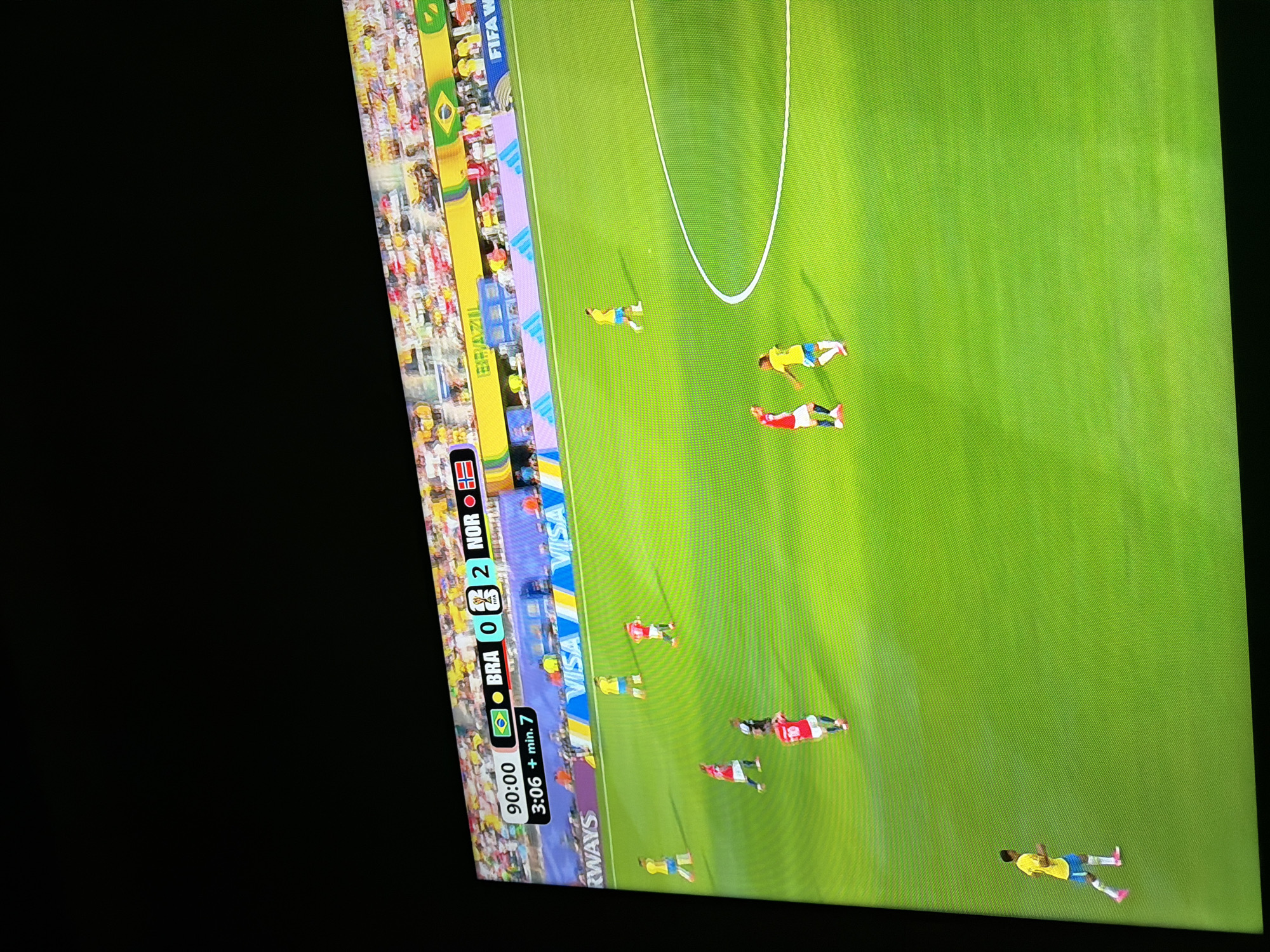 This is a photograph of a television screen displaying a soccer match. The score is Brazil 0, Norway 2. The game is in the 90th minute with an additional 7 minutes to be played. The stadium is filled with spectators wearing yellow and white, with some red visible. The field is green. The advertisement boards around the field display "VISA" multiple times and "RWAYS." There are several players on the field wearing yellow and blue uniforms (Brazil) and red uniforms (Norway). Two players in red uniforms are in the foreground, one with his arms raised. A player in a yellow and blue uniform is running towards the right of the frame. Another player in yellow and blue is near the center of the field. The photograph is taken from a slightly upward angle, and the screen itself has a visible texture from the pixels. The location provided, Ognianovo, Bulgaria, is not depicted or implied in the image; the image only shows a soccer match on a screen.