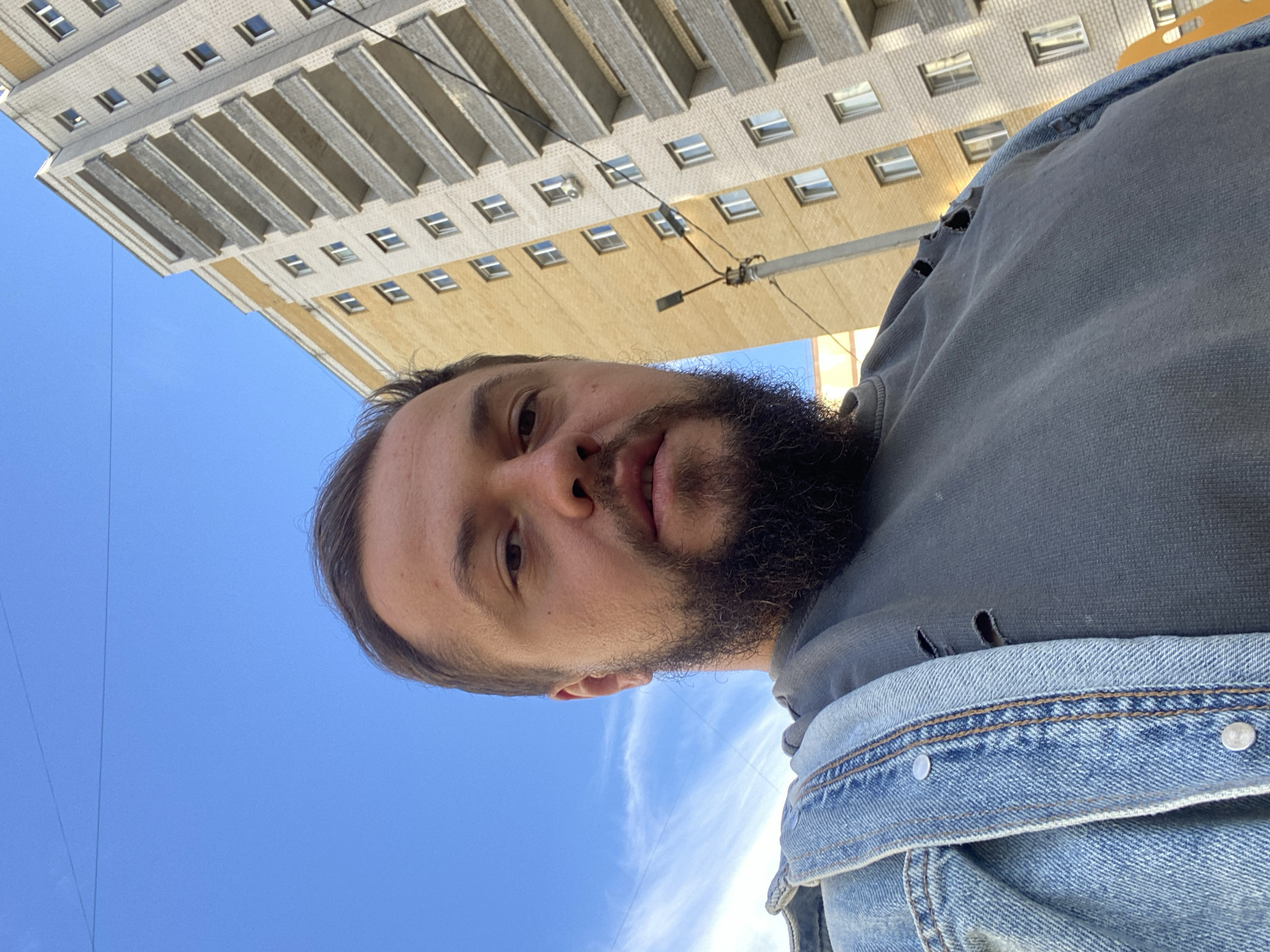 The image shows a close-up, upward-facing selfie of a man with a beard. He is wearing a denim jacket over a gray hoodie. The man is looking at the camera with a neutral expression.
The background of the image features a blue sky with some wispy clouds and a building with multiple windows. The building appears to be made of brick and has a beige facade. The angle of the shot suggests the photographer is looking up at the building. There are power lines visible in the sky.
The lighting indicates it is daytime, likely sunny due to the brightness. The overall impression is of an urban environment. No visible text is present in the image. The image is oriented in a way that the top of the man's head is towards the left and the bottom of the image is towards the right, meaning the image is rotated 90 degrees counter-clockwise.