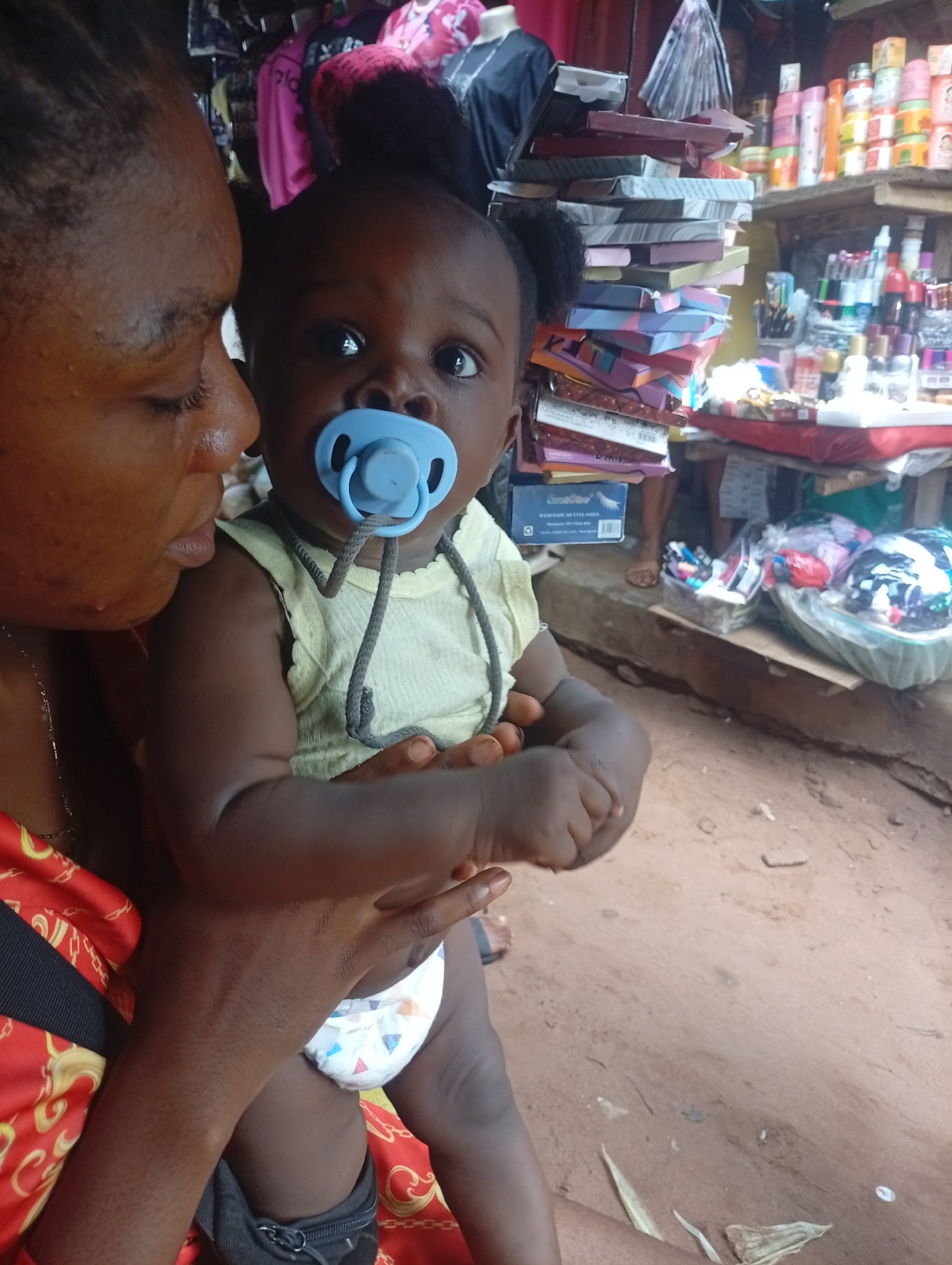 A person holds a baby in their arms. The person is looking down at the baby, who is facing forward and has a pacifier in their mouth. The baby is wearing a light-colored sleeveless shirt and a diaper. The person is wearing a red top with gold patterns. In the background, there are shelves stocked with various items, including cosmetics and stacked boxes. The ground appears to be made of dirt. The scene appears to be outdoors in a market setting.