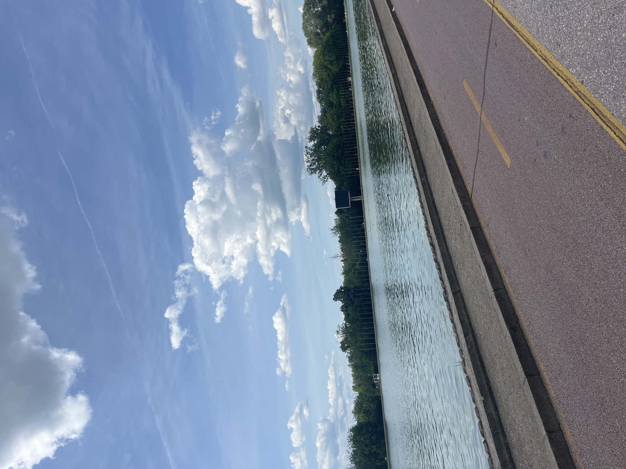 The image shows a body of water, likely a canal or river, adjacent to a pathway. The pathway is separated from the water by a concrete barrier. The pathway itself is made of red asphalt with a yellow dividing line. Beyond the water is a line of trees, likely bordering the opposite bank. The sky is blue with scattered white clouds. The image is likely taken in Plovdiv, Bulgaria, specifically near the Rowing Canal, though it's difficult to be 100% certain without other visual cues, as the waterway is similar. There are no people visible in the image. The scene is daytime and outdoors. The focus is on the waterway and pathway.