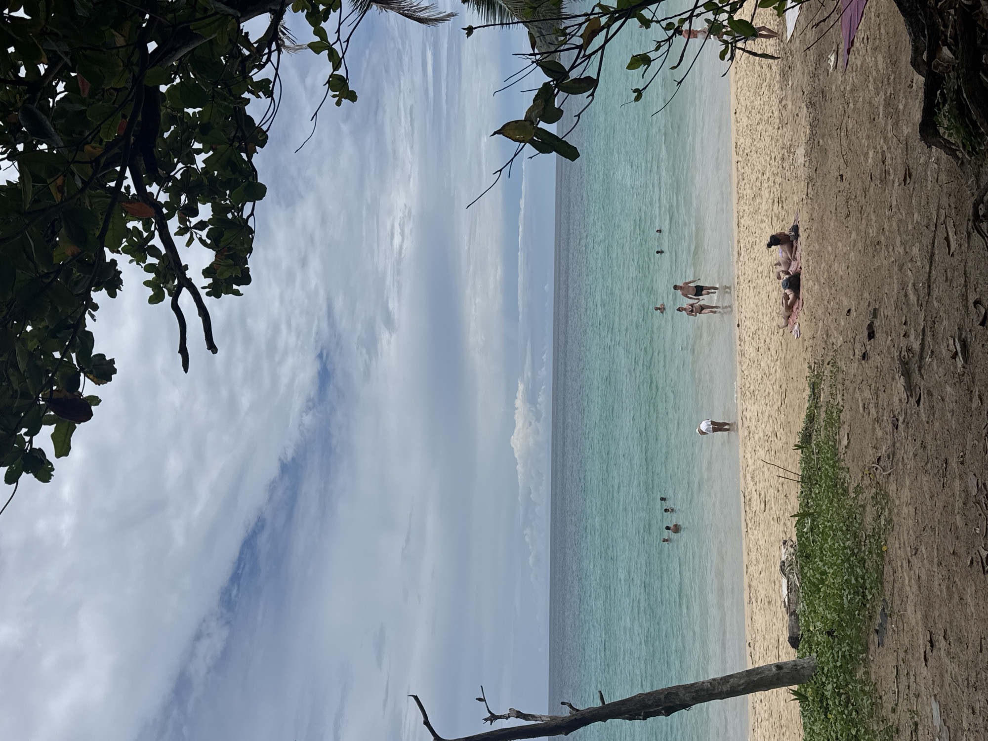The image captures a beach scene in Rawai, Thailand. The foreground is dominated by sandy terrain with fallen leaves and tree roots, bordered by lush green vegetation on the left. A weathered tree trunk stands prominently on the left side of the beach. In the middle ground, the sandy beach transitions into the clear, turquoise waters of the ocean. Several people are visible in the water, with some wading near the shore and others further out. On the beach, two individuals are seen lounging on towels, and another person stands near the water's edge. The background features the vast expanse of the ocean meeting a cloudy sky with distinct cloud formations. The overall atmosphere suggests a relaxed day at the beach.
