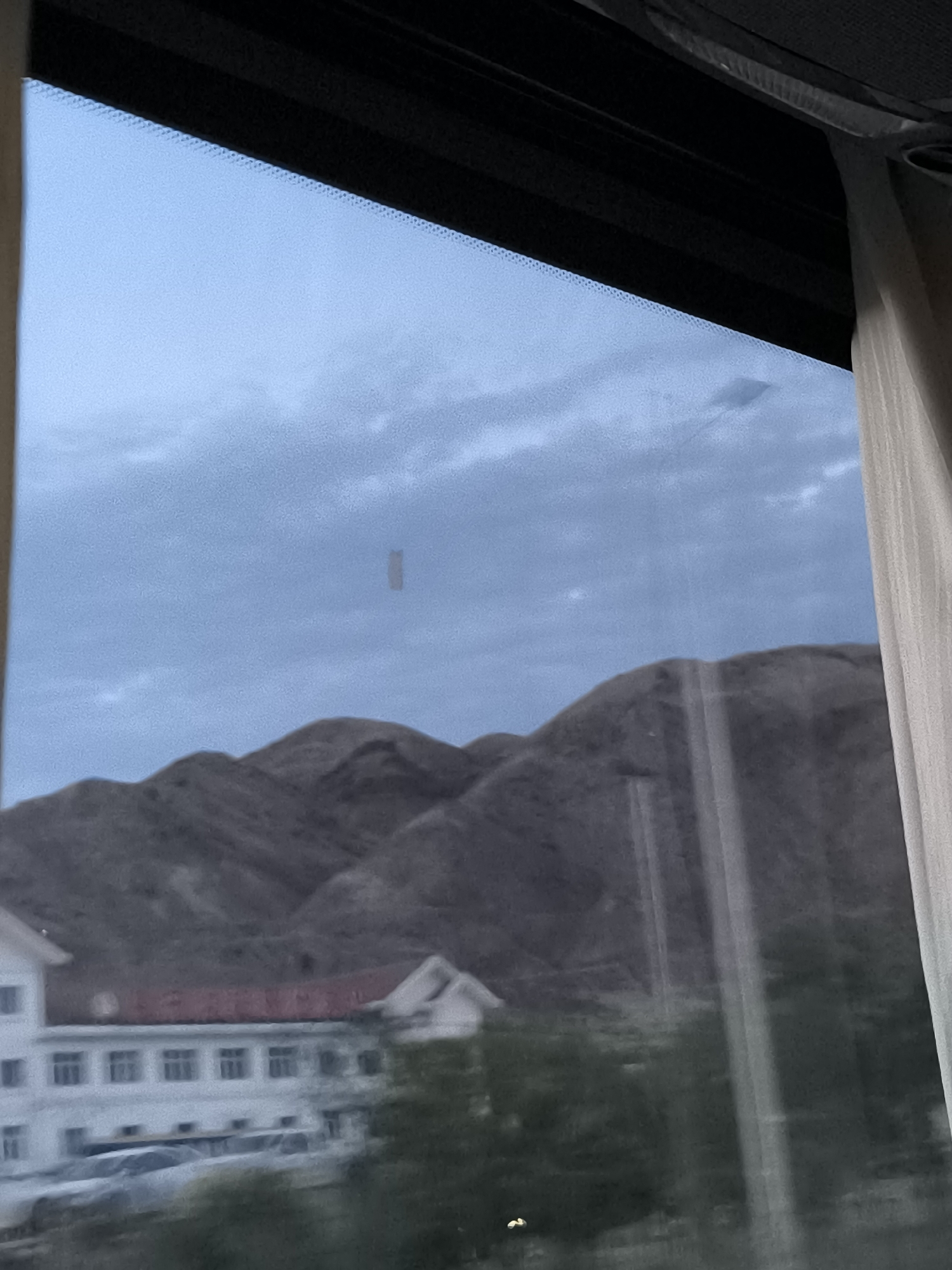 The image captures a view through a window of a building, likely a bus or train, at 果子沟牧场, China. In the foreground, a white curtain is visible on the right, and the top edge of the window frame is dark. The window looks out onto a landscape with rolling, barren mountains under a hazy, overcast sky. The sky has a light blue hue with scattered clouds. A faint, rectangular, translucent object is visible in the sky. Below the mountains, there is a building complex with white walls and a reddish-brown roof, and trees are sparsely scattered in the middle ground. The overall scene appears to be a rural or semi-rural environment, possibly during the early morning or late evening due to the dim lighting.