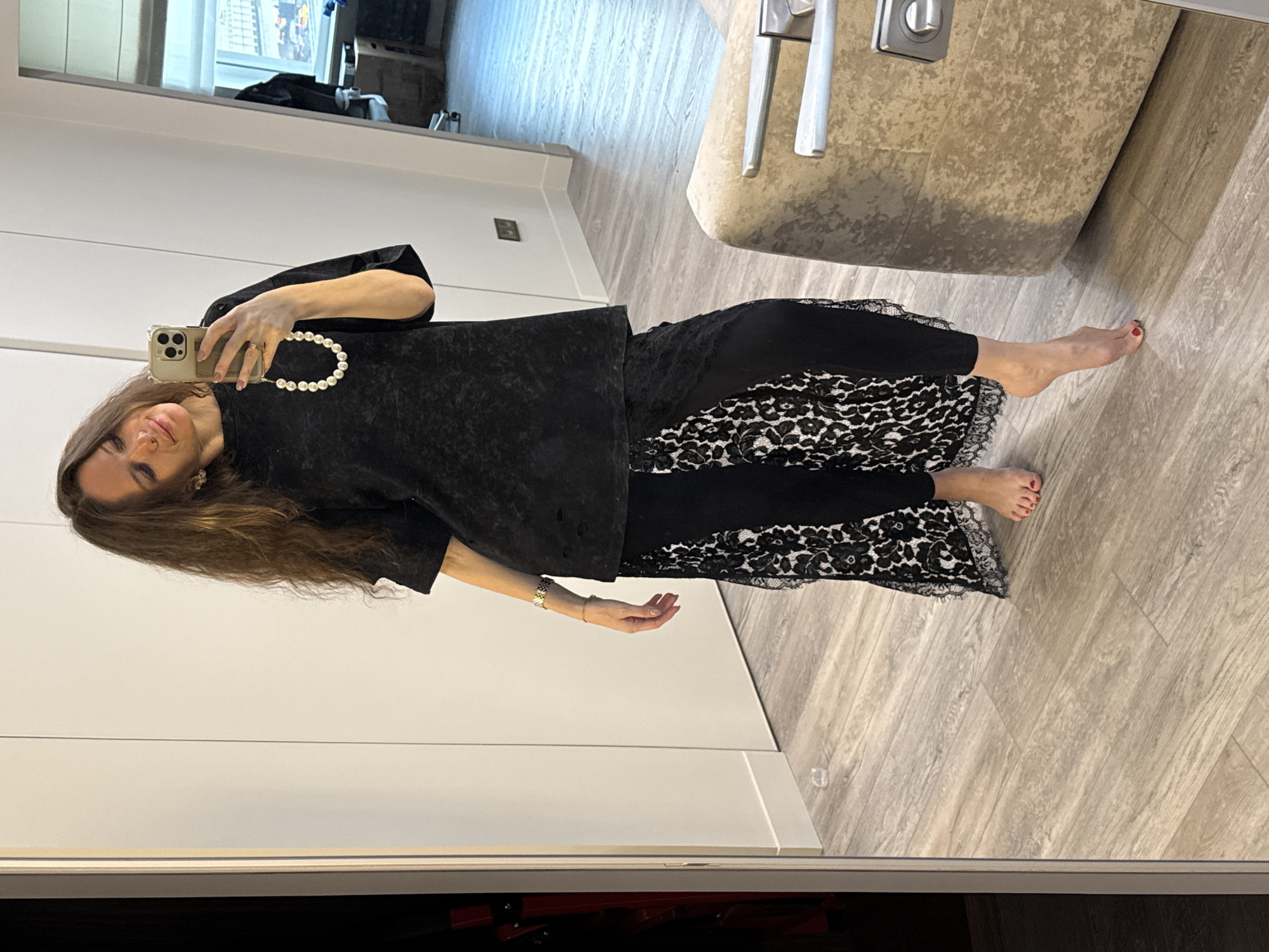 A woman is taking a selfie in a mirror. She is wearing a black top, a black skirt with a lace overlay, and a pearl necklace. She has long brown hair. The mirror is in a room with wood floors and white walls. A beige chair is visible in the background. The location appears to be an interior, possibly a home or apartment. There is no explicit indication that this image was taken in Dolgoprudny, Russia.