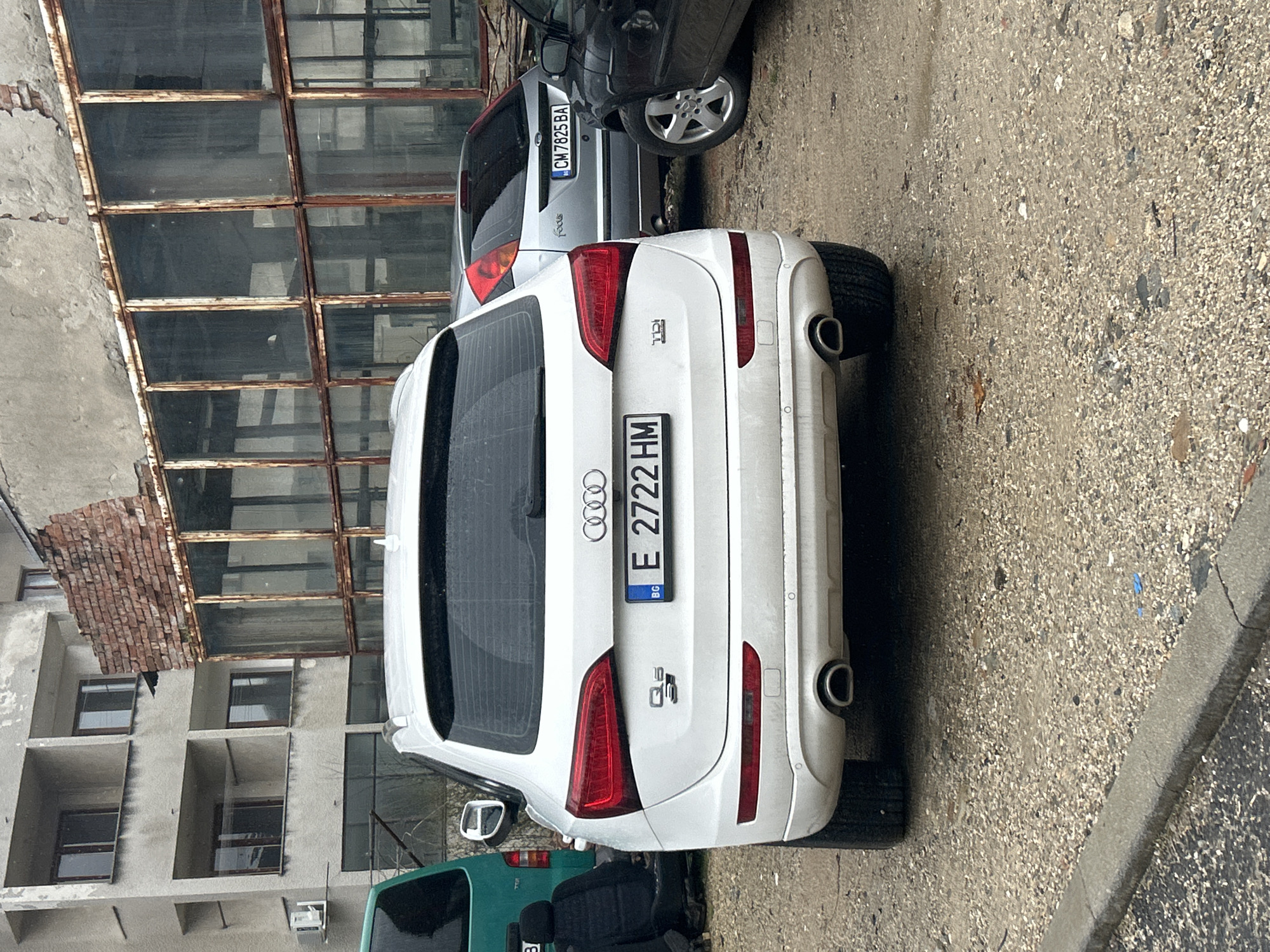 The image shows a white Audi Q5 parked on a street in Zlatograd, Bulgaria. The license plate reads "E 2722 HM". A silver Ford Focus with license plate "CM 7825 BA" is parked nearby. The environment includes an old, rundown building with rusty window frames and a partially demolished brick wall. The street surface appears to be paved with small stones or gravel. Another green car is partially visible behind the Audi. No people are visible in the image.
