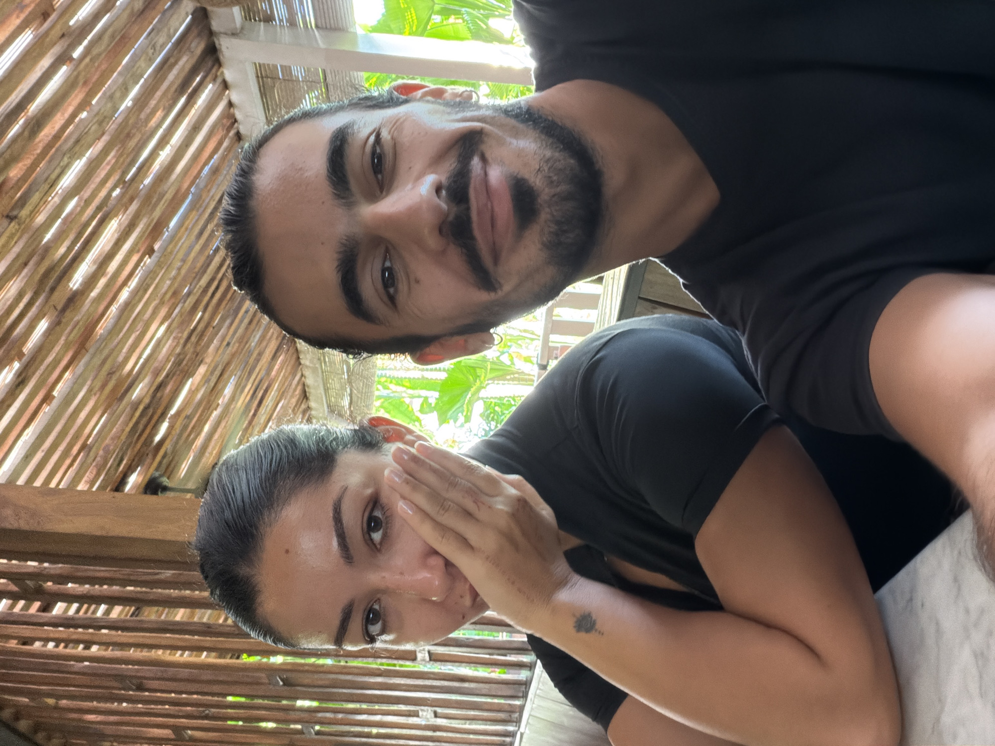 A selfie captures a man and a woman in what appears to be an outdoor or semi-outdoor setting, potentially in North Kuta, Indonesia. The man, positioned at the top of the frame, has dark hair, a beard, and a mustache, wearing a black t-shirt. He is smiling slightly. The woman is located lower in the frame with dark hair pulled back, and she is wearing a black t-shirt. Her left hand is raised near her face, partially obscuring it. A small tattoo is visible on her left forearm. The background includes a partially constructed wooden structure and green foliage, suggesting a tropical environment.