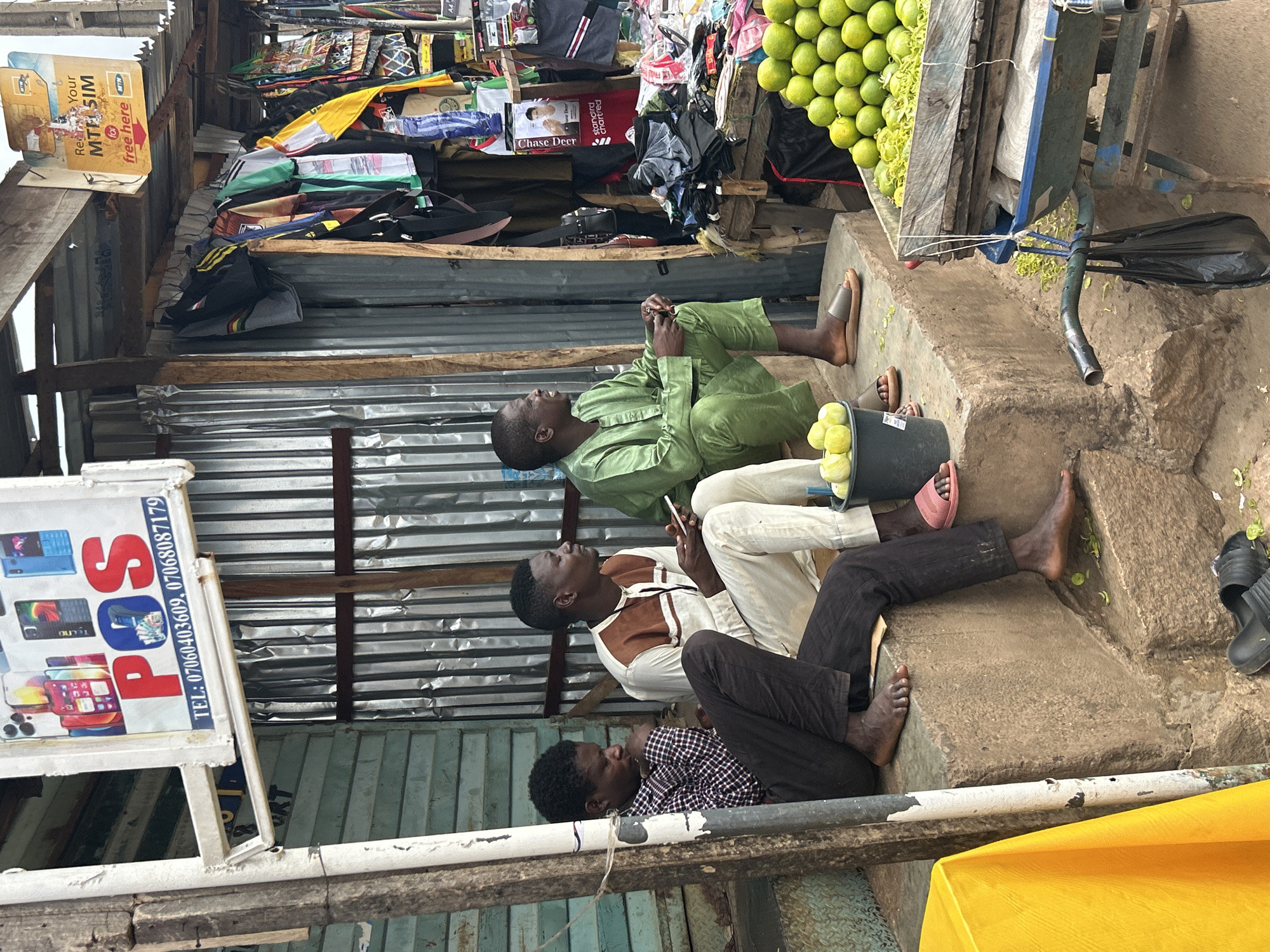 This vertical image depicts an outdoor market or street scene in Jos, Nigeria, showcasing three men resting amidst commercial activities. The setting is characterized by makeshift structures of corrugated metal sheets and wooden beams, with rough concrete steps and ground serving as seating and display areas.
In the immediate foreground, to the bottom right, a man in a checkered shirt and dark trousers sits on a concrete step, appearing to be resting or observing his surroundings. Next to him, a man wearing a white top with brown accents and light-colored trousers is engrossed in a white smartphone, looking upwards. A black bucket filled with green citrus fruits (limes or small lemons) rests near his bare feet. Further back and to the left, another man in a light green traditional outfit, possibly an agbada, is also looking upwards, his hands clasped or holding an unseen object. He wears sandals.
The background reveals a bustling market environment. To the right, a large pile of green limes or small lemons is prominently displayed on a cart or stall. Above the men, various merchandise including bags, clothing items, and other goods are stacked on shelves.
Several signs add to the commercial ambiance: a yellow sign prominently advertises "Re Your MTN SIM free here," indicating a mobile network service. A white sign with red lettering displays "POS" and contact numbers "TEL: 07060403609, 07068087179," accompanied by images of smartphones, including a "TECNO" device, suggesting an electronics or financial service point. Other partially visible text includes "Chase Deer" and "standard chartred."
The bright, even illumination indicates it is daytime. The scene conveys a relaxed atmosphere where people are either waiting, resting, or observing their environment in a busy, informal market setting.