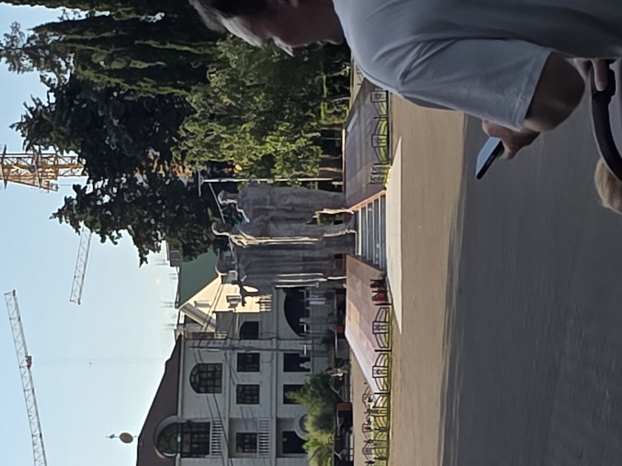 A medium shot captures a group of people and a monument in Sochi, Russia, under a clear sky. In the foreground, a person in a white t-shirt is seen from behind, holding a black smartphone. To the left of this person, a blonde child's head is visible, partially obscured by a stroller. The middle ground features a large, stone monument depicting several figures standing together. The monument is elevated on a tiered base with a sloped ramp leading up to it. A decorative metal fence encloses the area around the monument. Behind the monument, several buildings are visible, including a white building with arched windows and a green-roofed building under construction. In the background, tall trees and two construction cranes tower over the scene, one yellow and one white, suggesting ongoing development in the area. The ground appears to be paved.